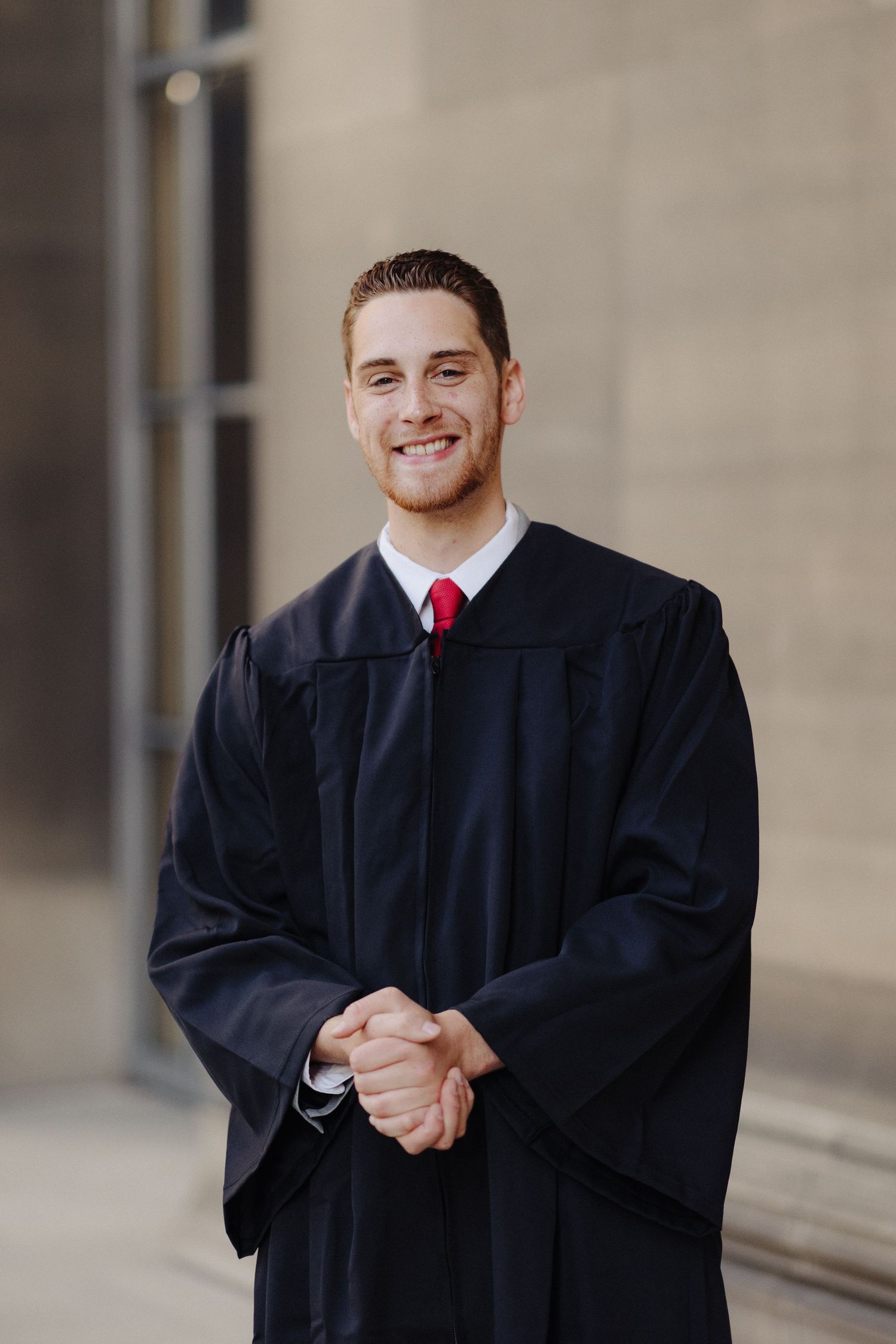 Graduate in dark academic regalia smiles while clasping hands in formal portrait.