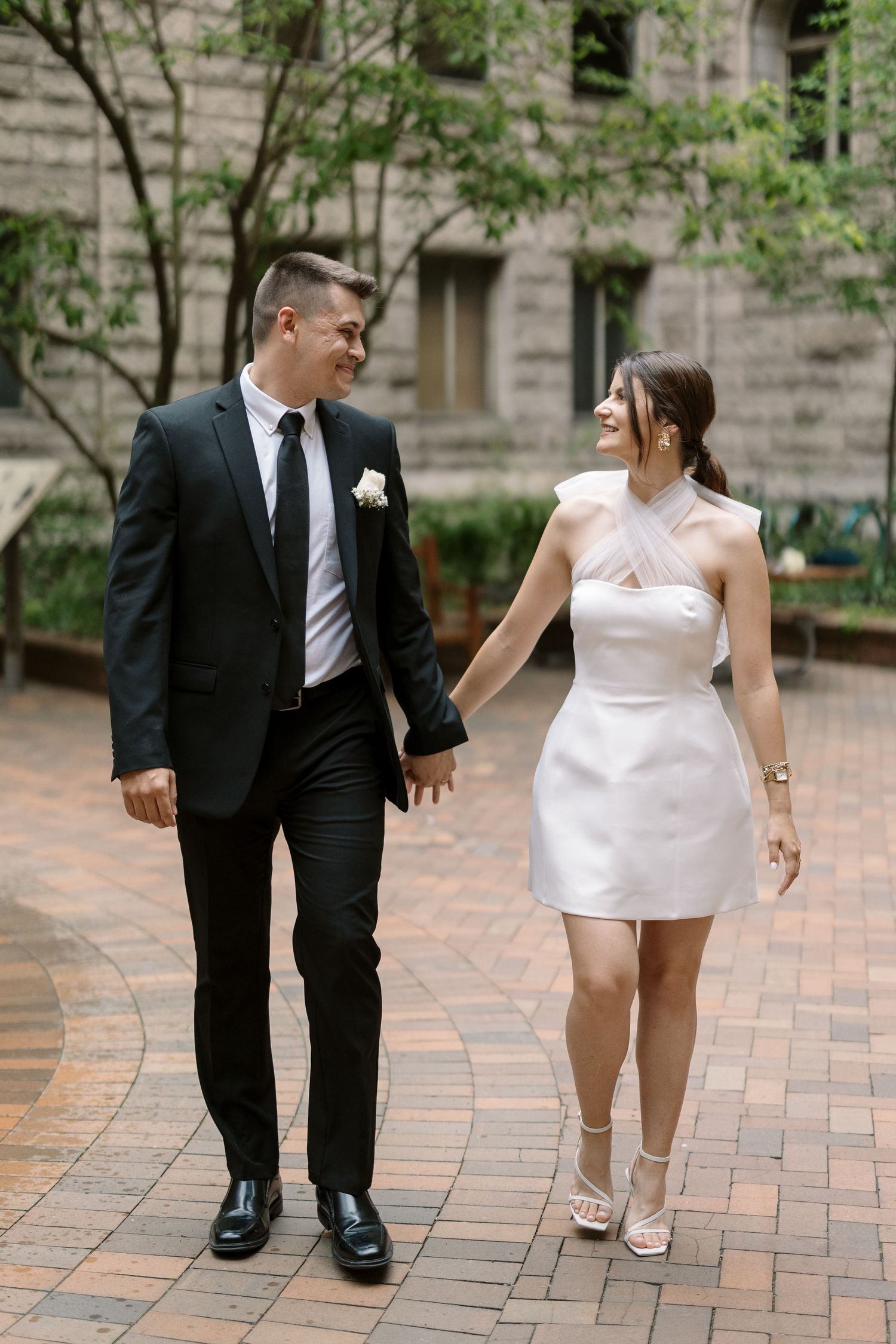 A couple walks together while holding hands in a courtyard with stone architecture.