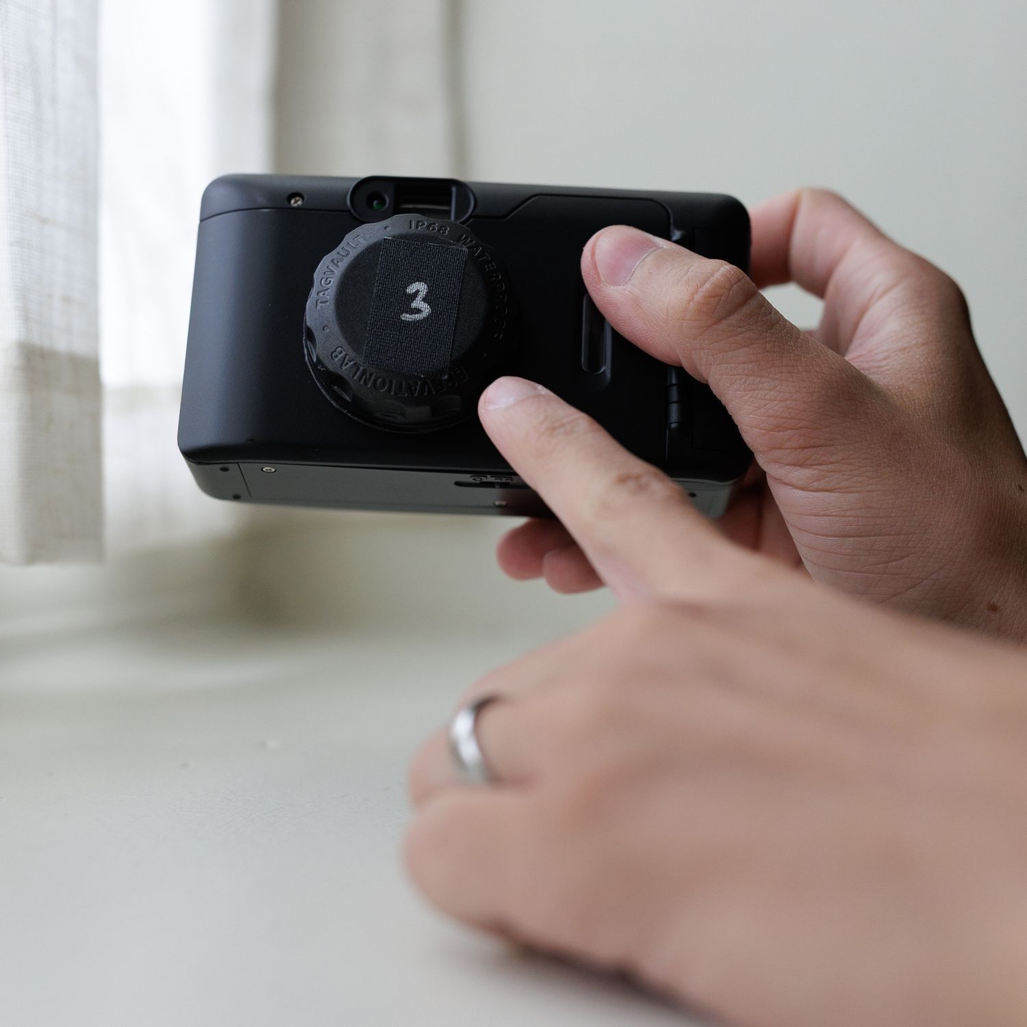 Hand holding a black camera or electronic device with a number 3 visible on its surface in a brightly lit interior.