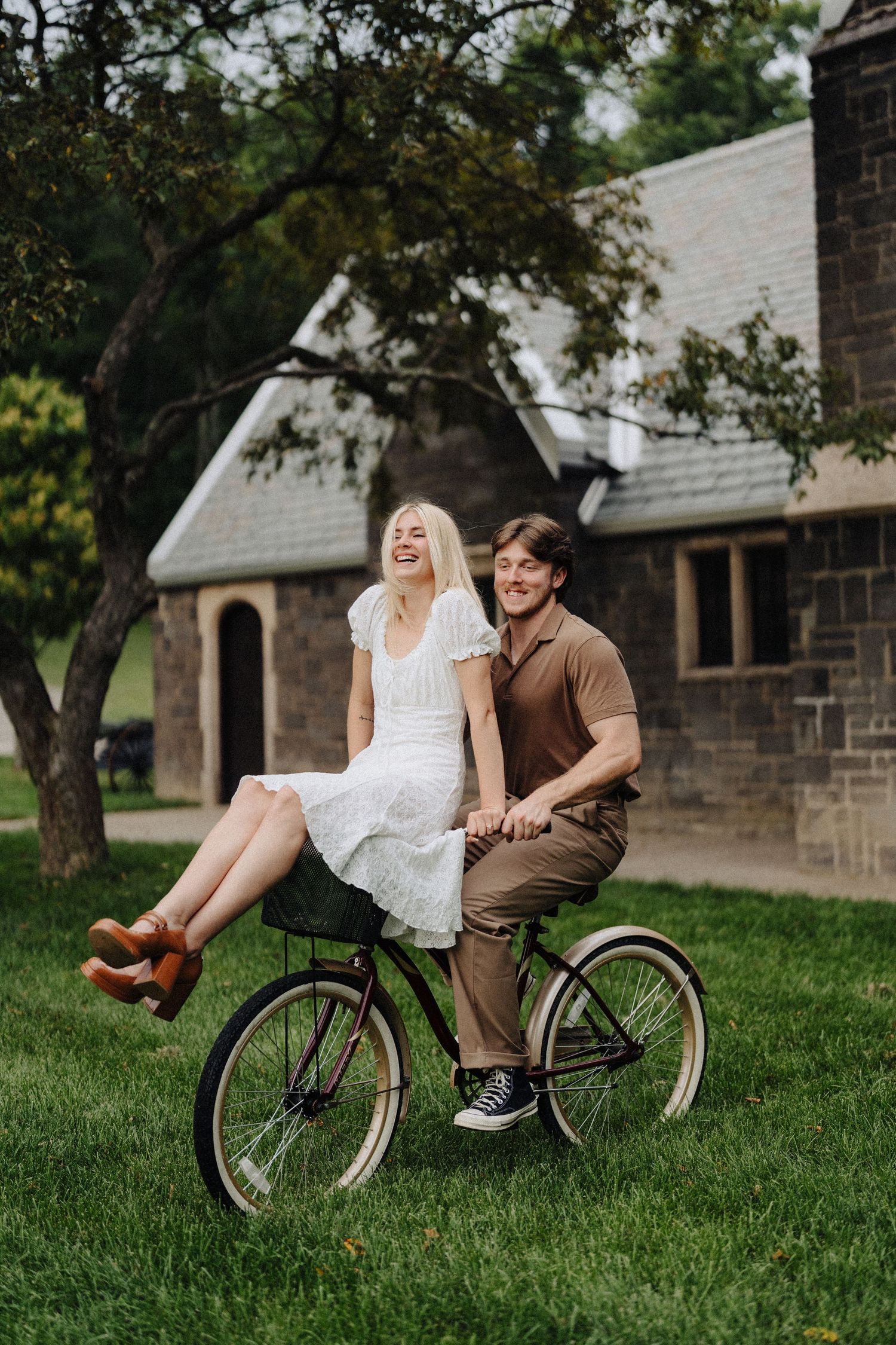 A playful couple enjoys a bike ride on the lawn of a stone church.