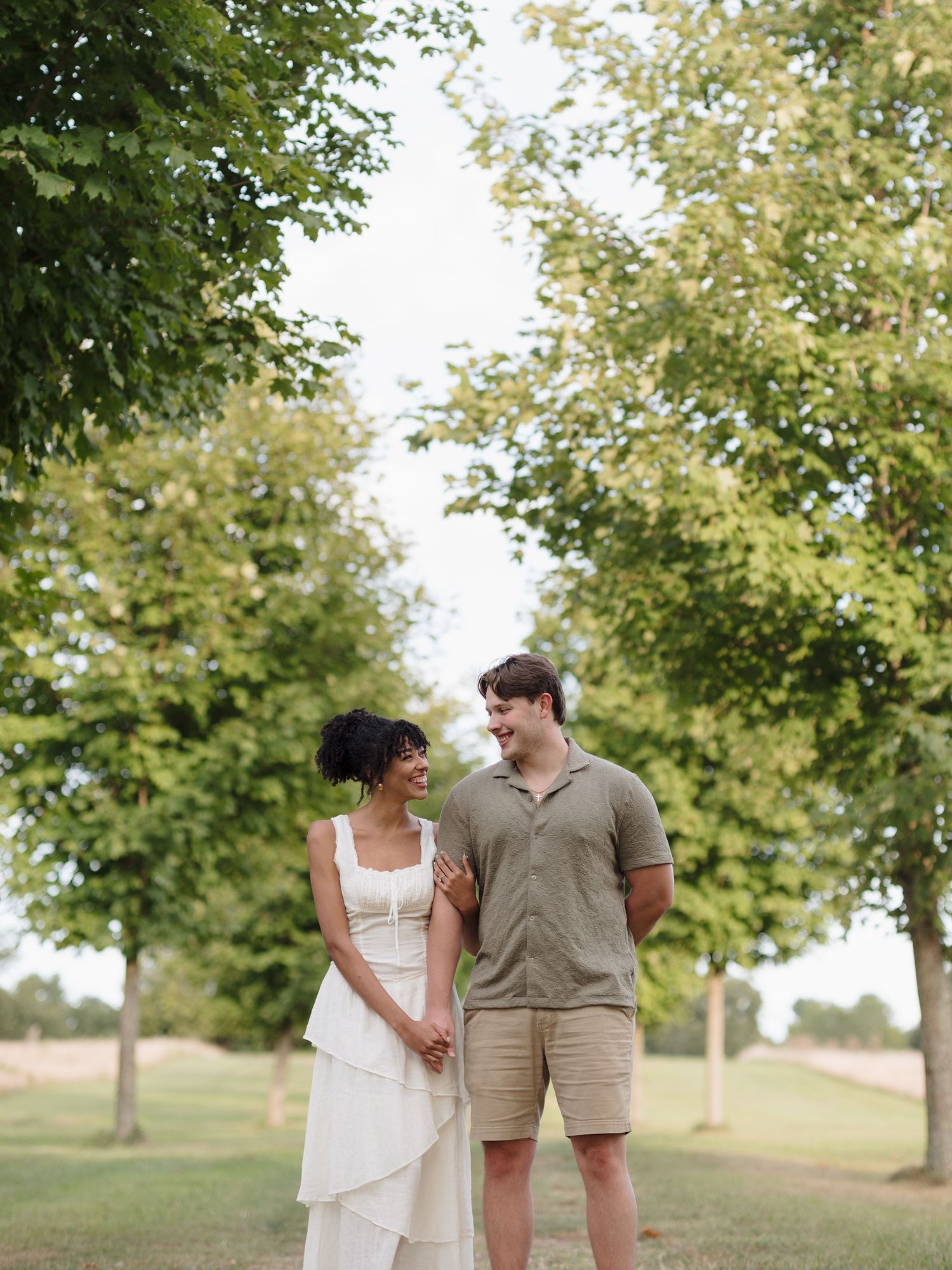 A couple in casual attire walks and laughs together along a tree-lined path on a bright summer day.