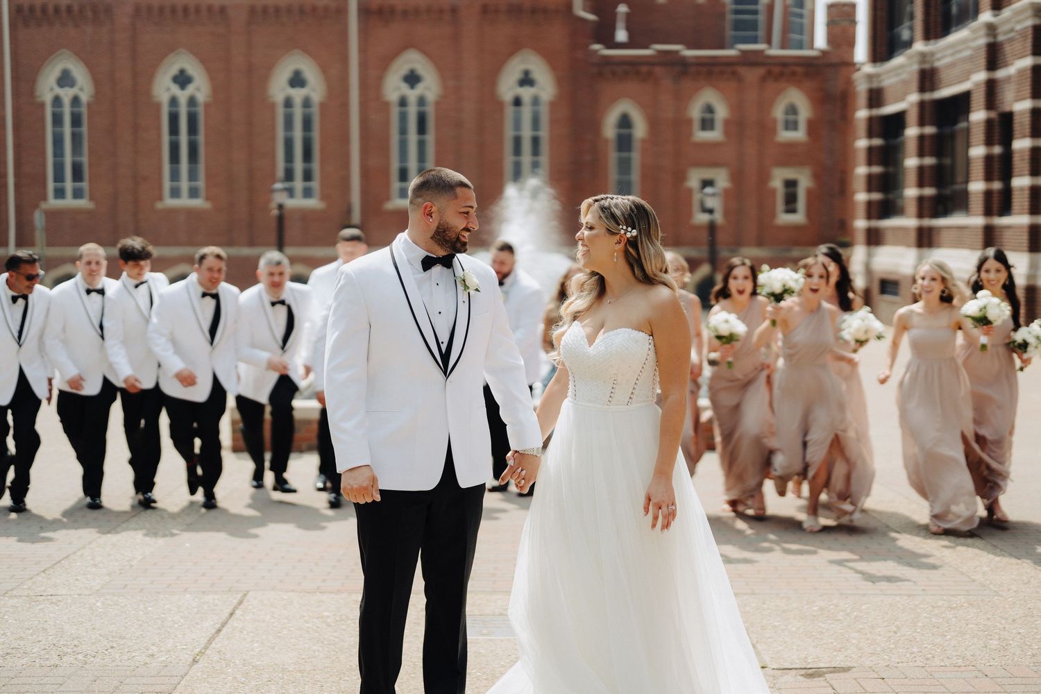 Wedding couple walks with bridal party in white and neutral colors behind brick church building.