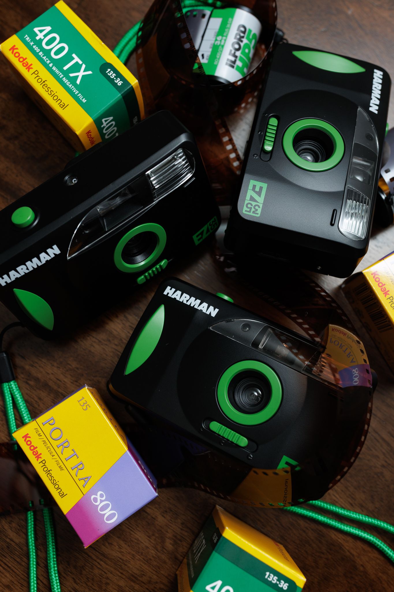 Several vintage Harman film cameras with green accents sit alongside rolls of Kodak and Fuji film on a wooden surface.