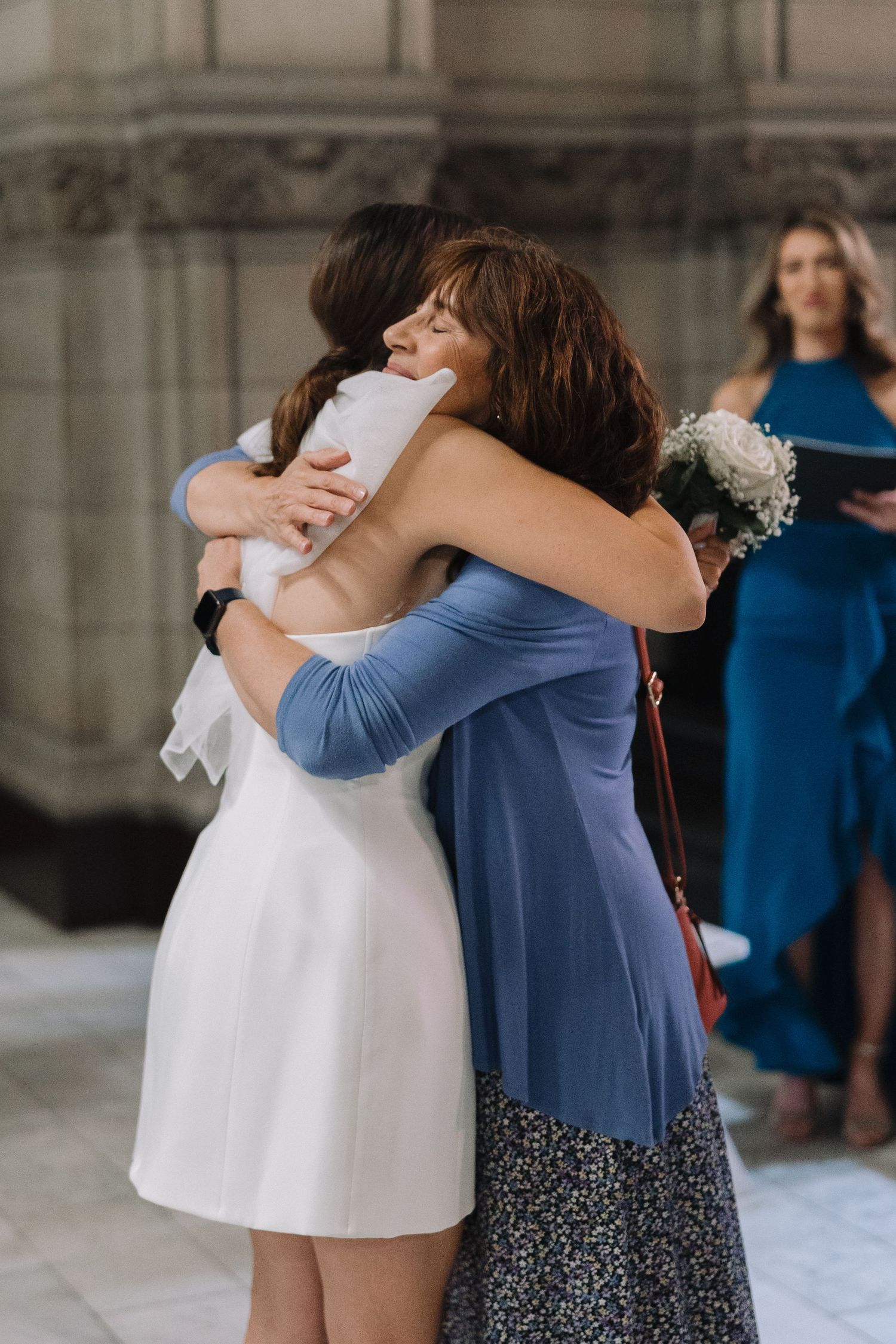 Emotional embrace during wedding ceremony with attendant in blue dress looking on.
