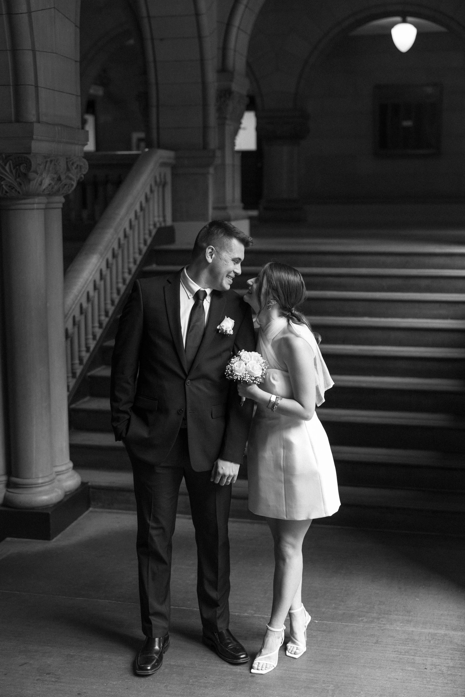 A couple in wedding attire share an intimate moment by a grand stone staircase in a historic building.