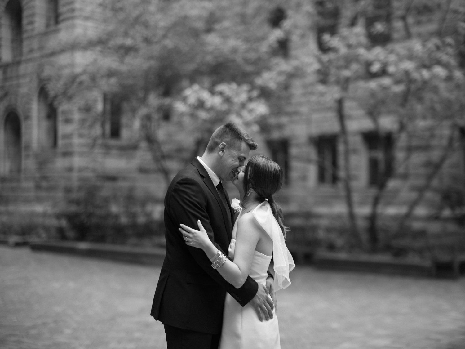 A black and white romantic wedding photo showing a couple embracing outdoors near a stone building.