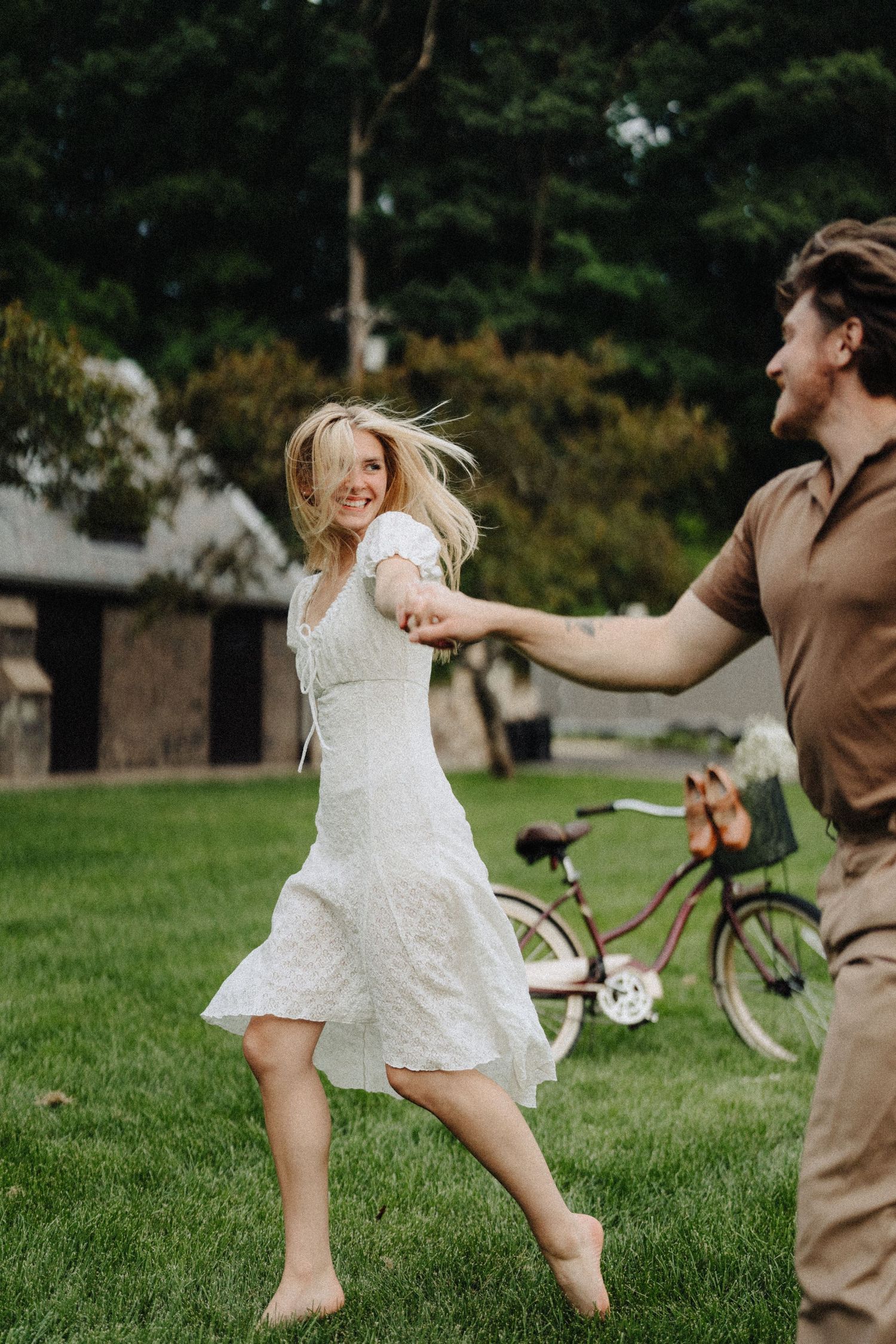 Dancing barefoot on green grass with a vintage bicycle in the background near stone buildings.