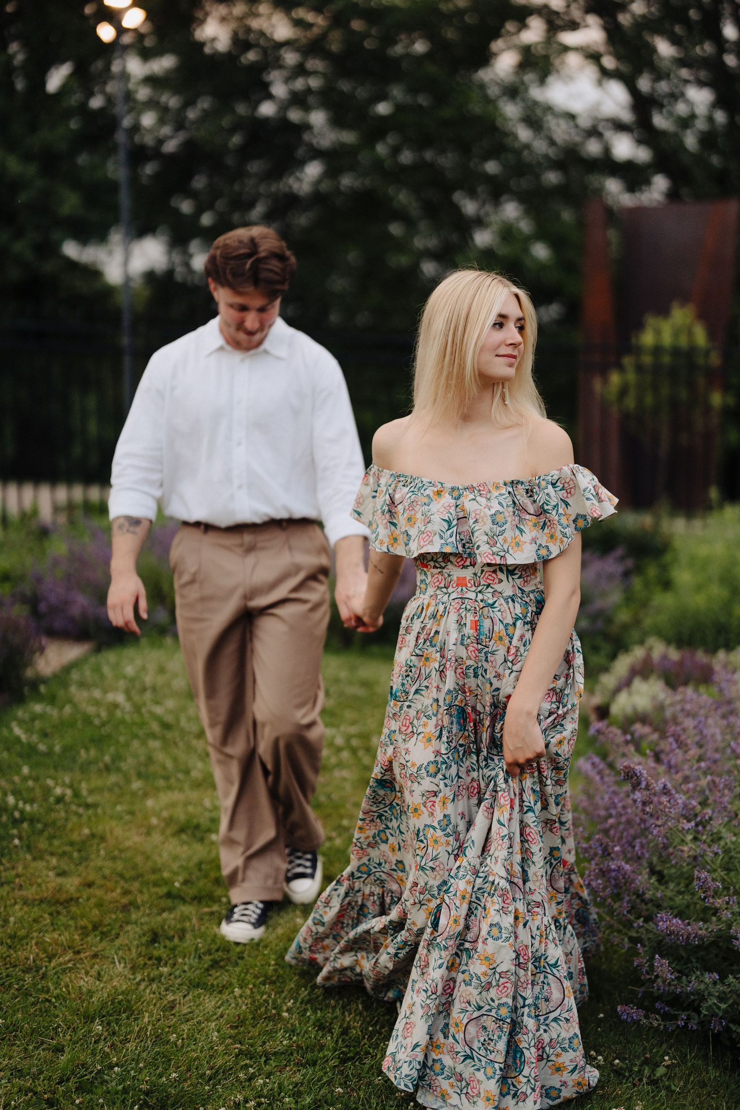 Couple strolls through blooming garden with floral dress and casual attire.