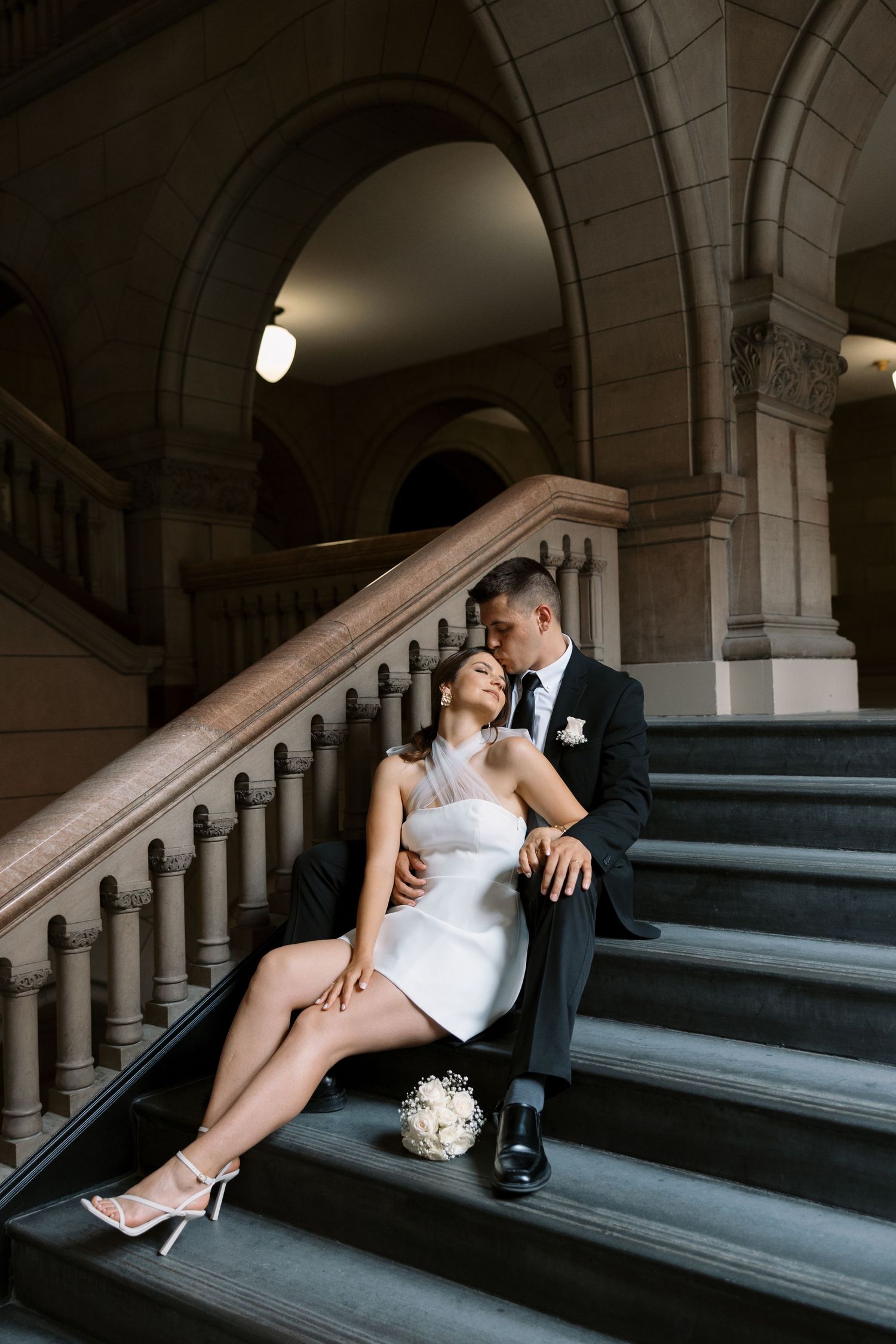 A romantic wedding photo of a couple posing on elegant stone stairs with vaulted architecture.