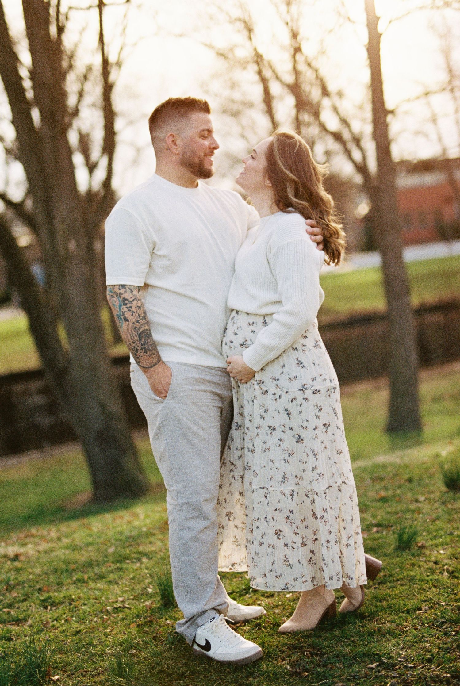 Romantic couple in white outfits share tender moments during outdoor photoshoot at sunset in a grassy park setting.