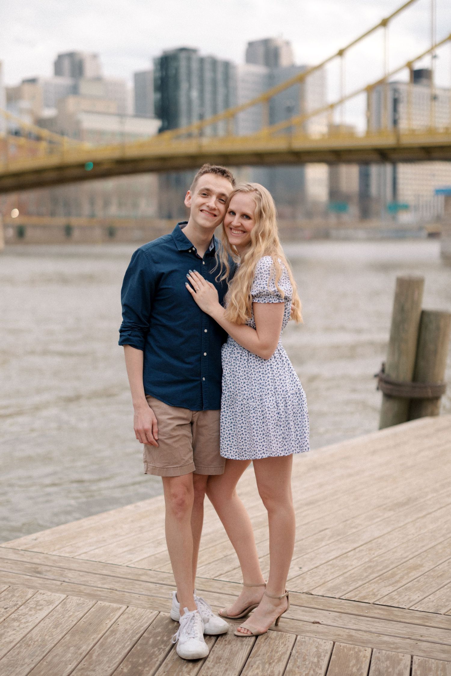 A couple poses together on a wooden pier with a yellow bridge and city skyline behind them.