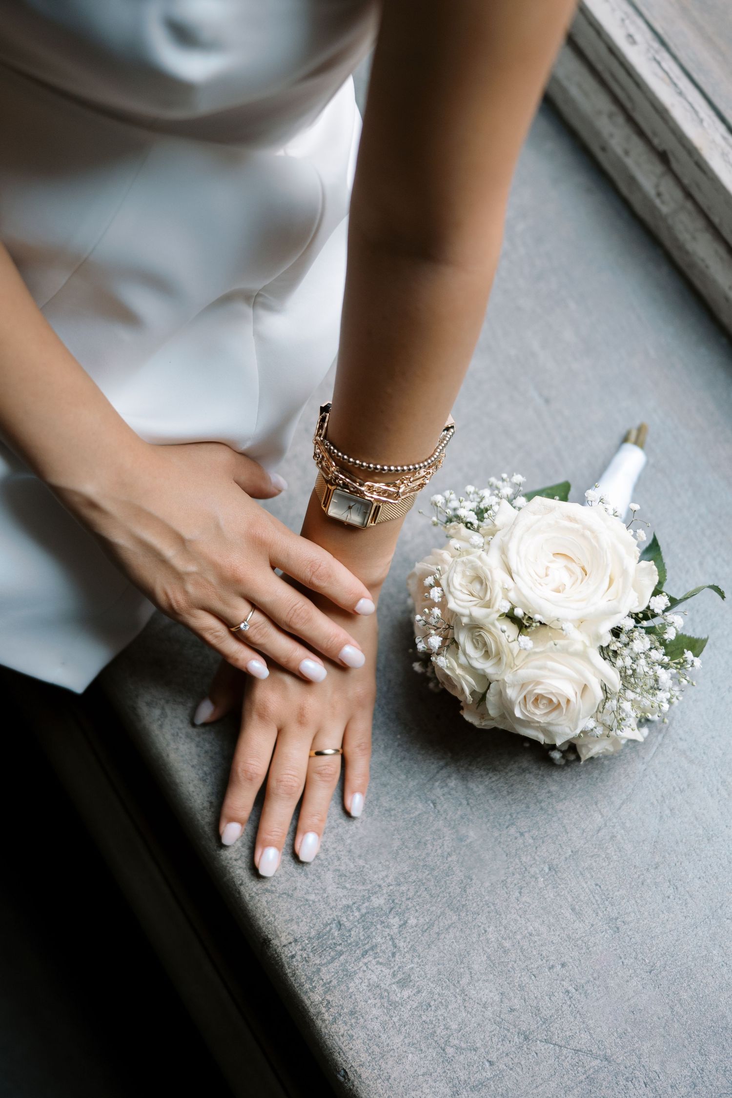 Wedding details featuring a white bouquet and jewelry on a bride's hands near a window.