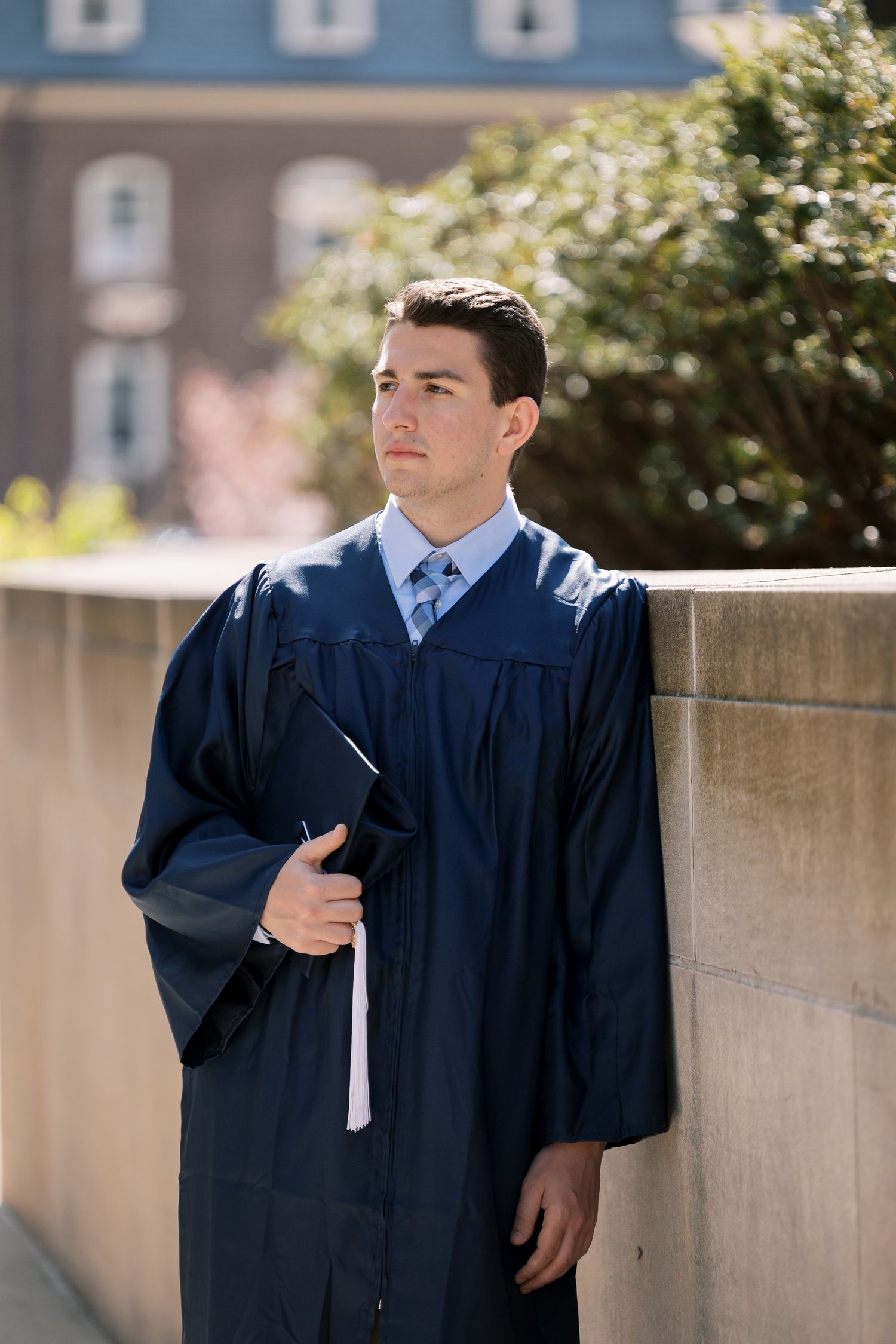 Graduate in navy blue cap and gown holding diploma stands thoughtfully against concrete wall on sunny day.