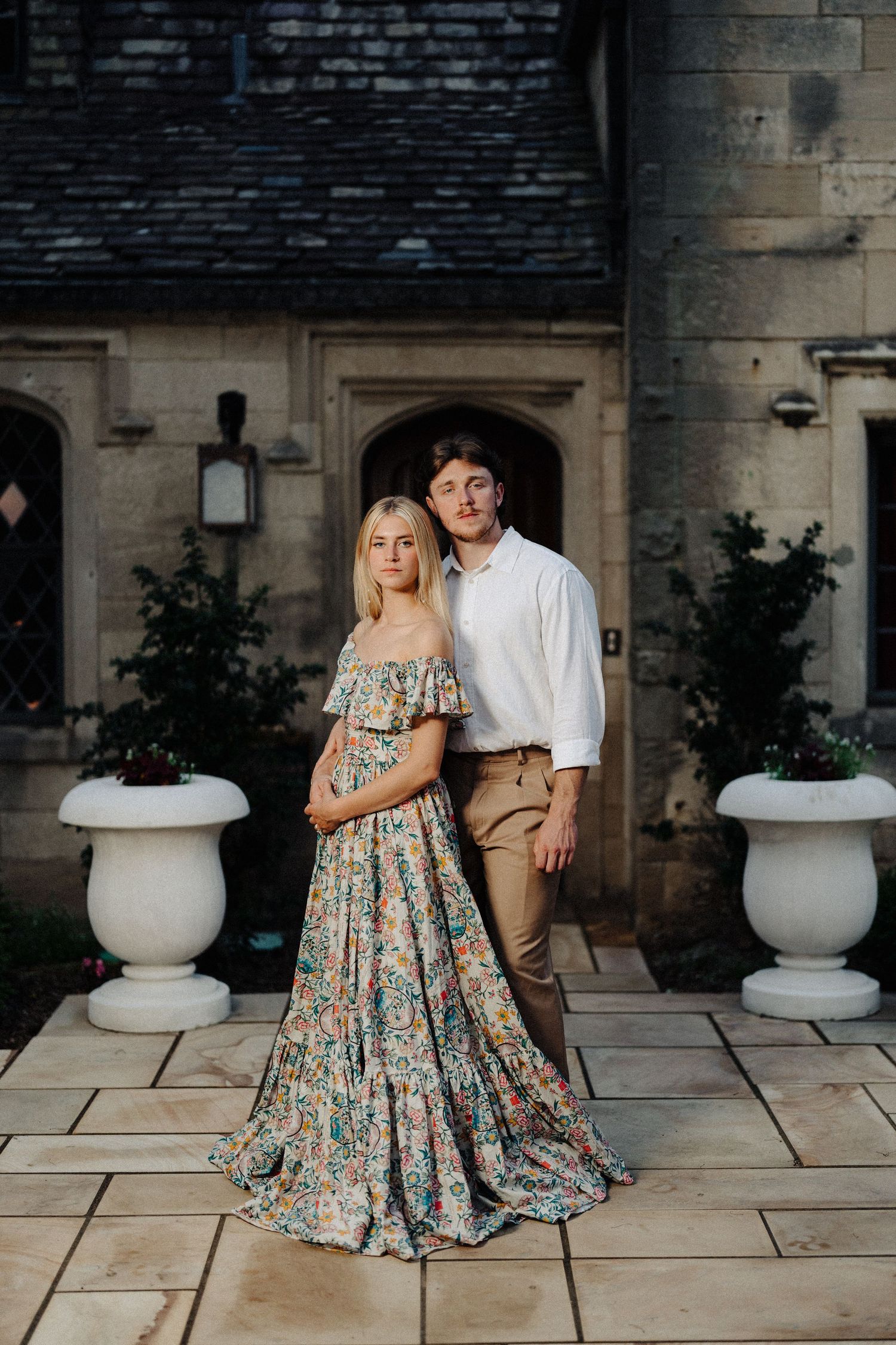 Elegant couple poses in formal attire against stone architecture with decorative urns.