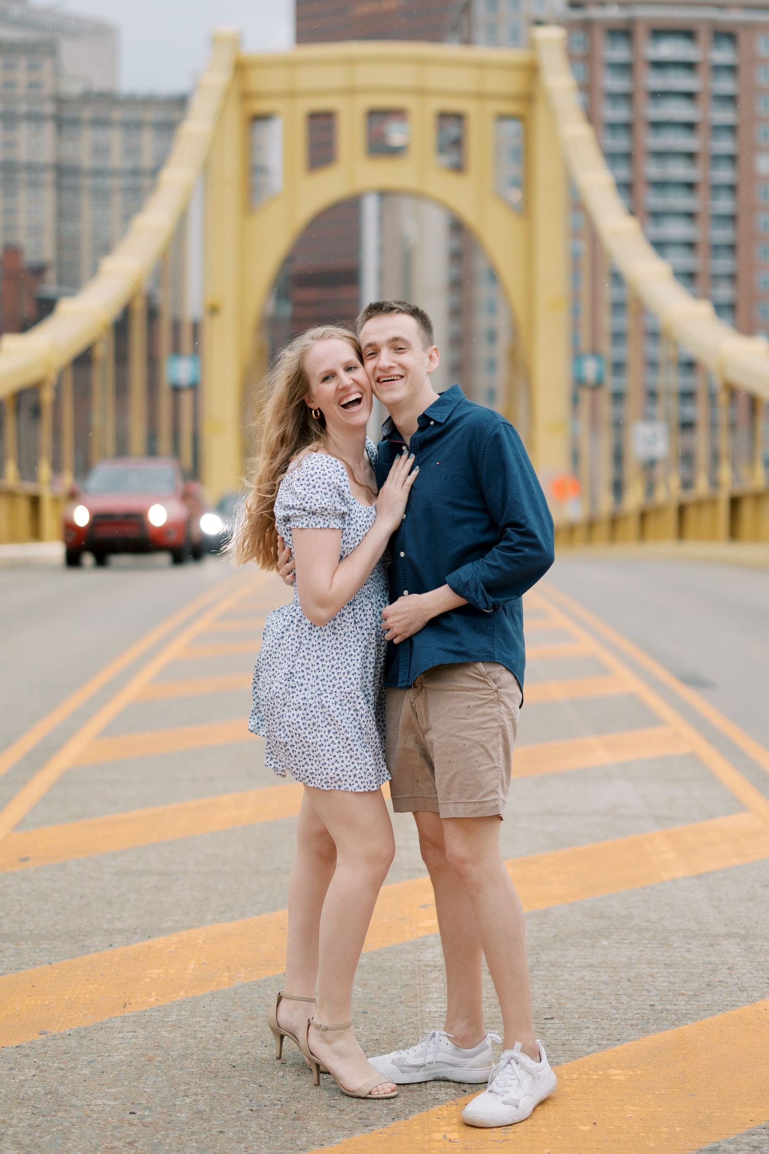 A couple shares a tender moment on a yellow bridge with city buildings in the background.