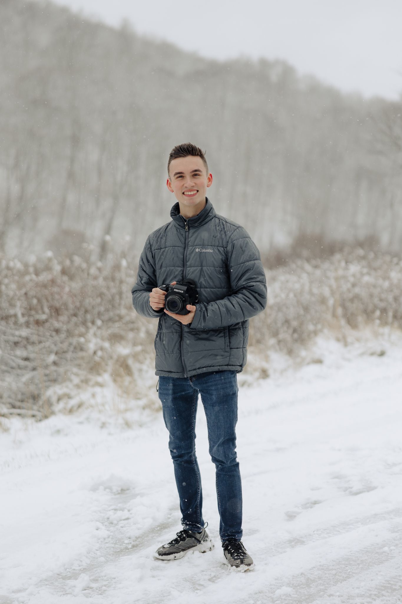 A photographer in a gray winter jacket holds a camera while standing in snowy winter landscape.