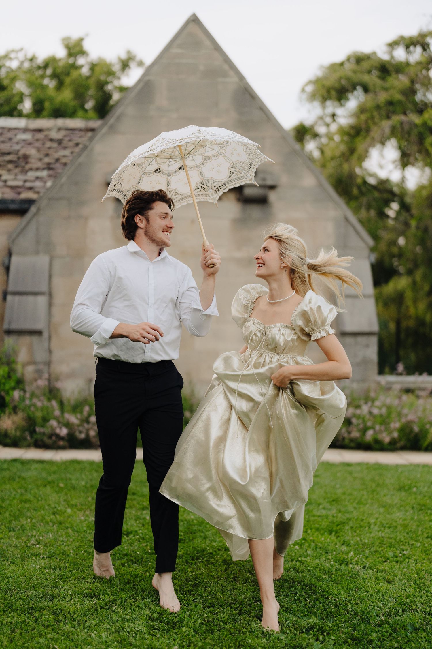 Playful couple running with lace umbrella on church lawn in vintage-inspired photoshoot.