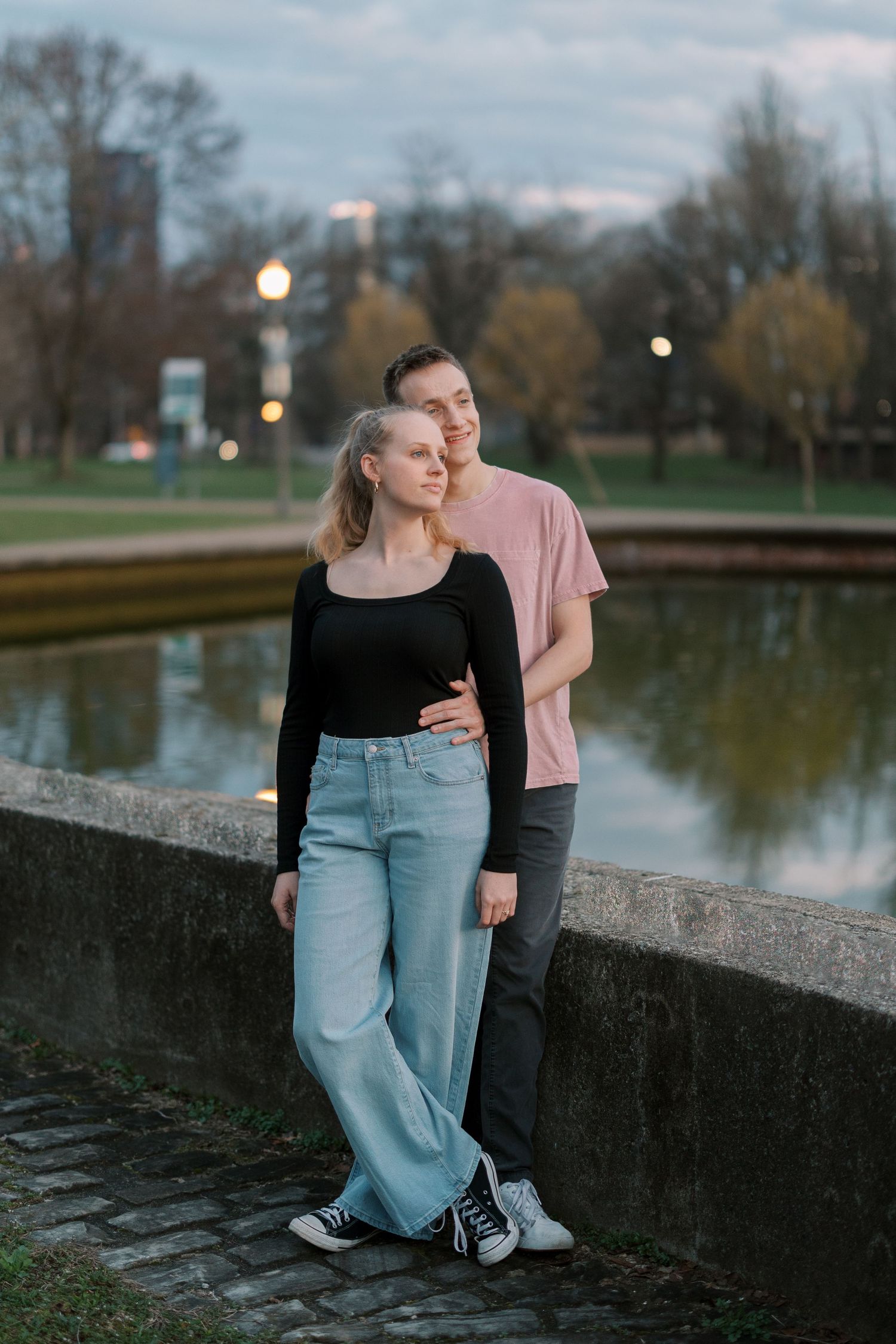 A couple stands together by a pond at dusk with trees and city lights in the background.
