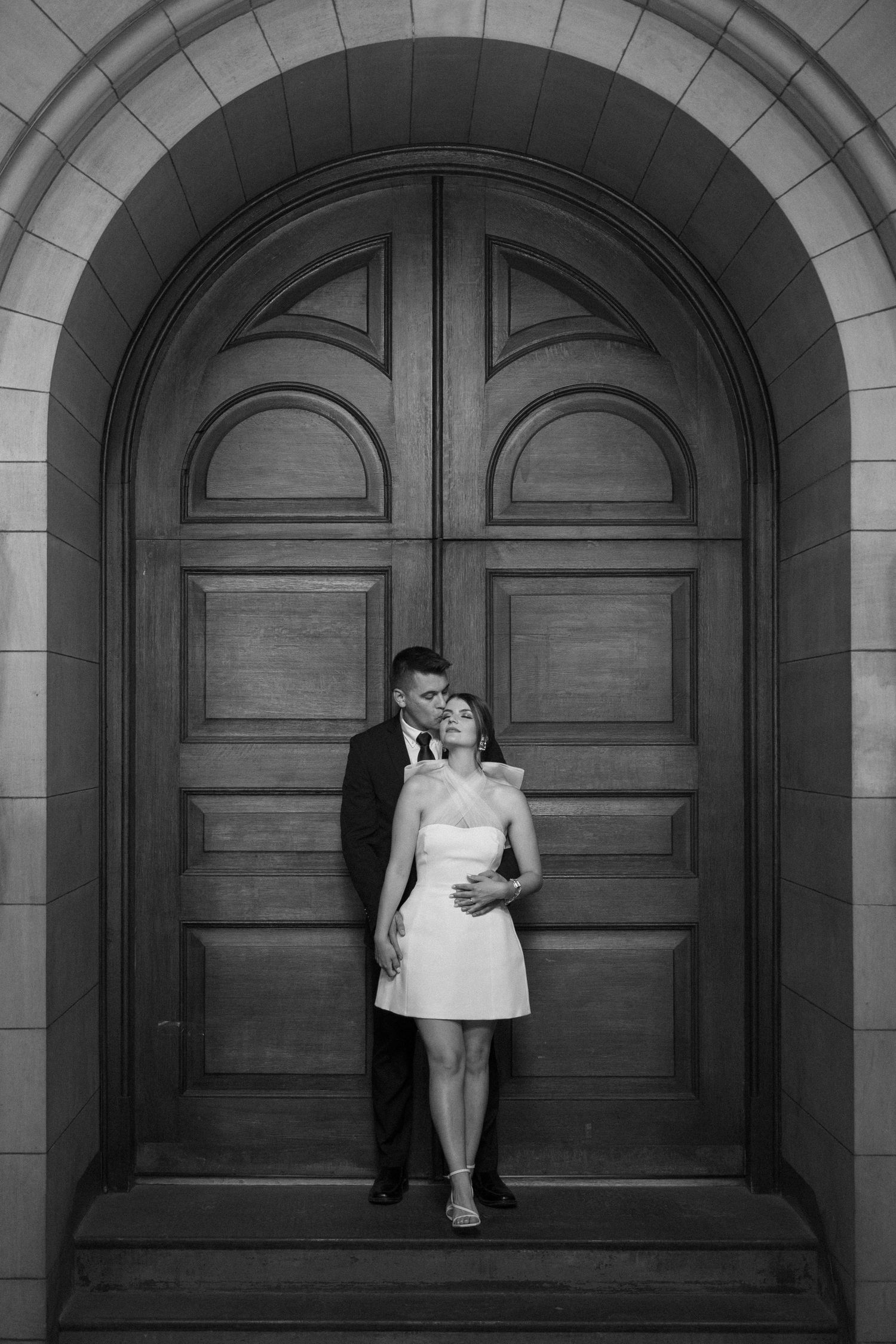 A romantic black and white photo of a couple standing against an elegant arched wooden door.