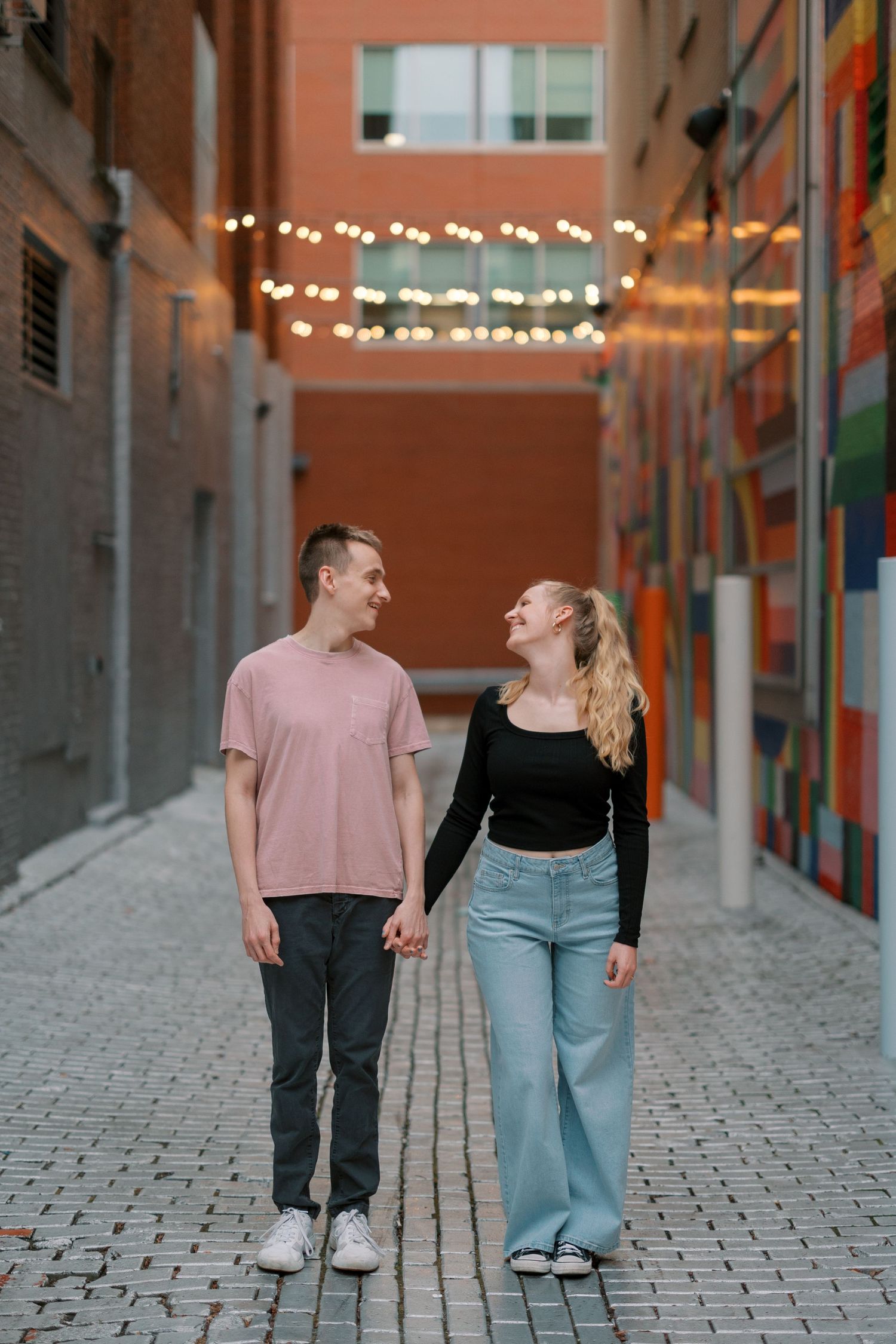 A couple walking together under string lights in an alley with a colorful mural.