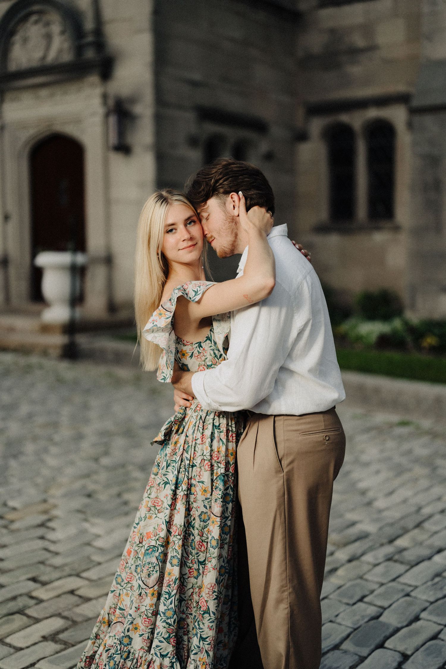 A couple shares an intimate moment in formal wear outside a historic stone building.