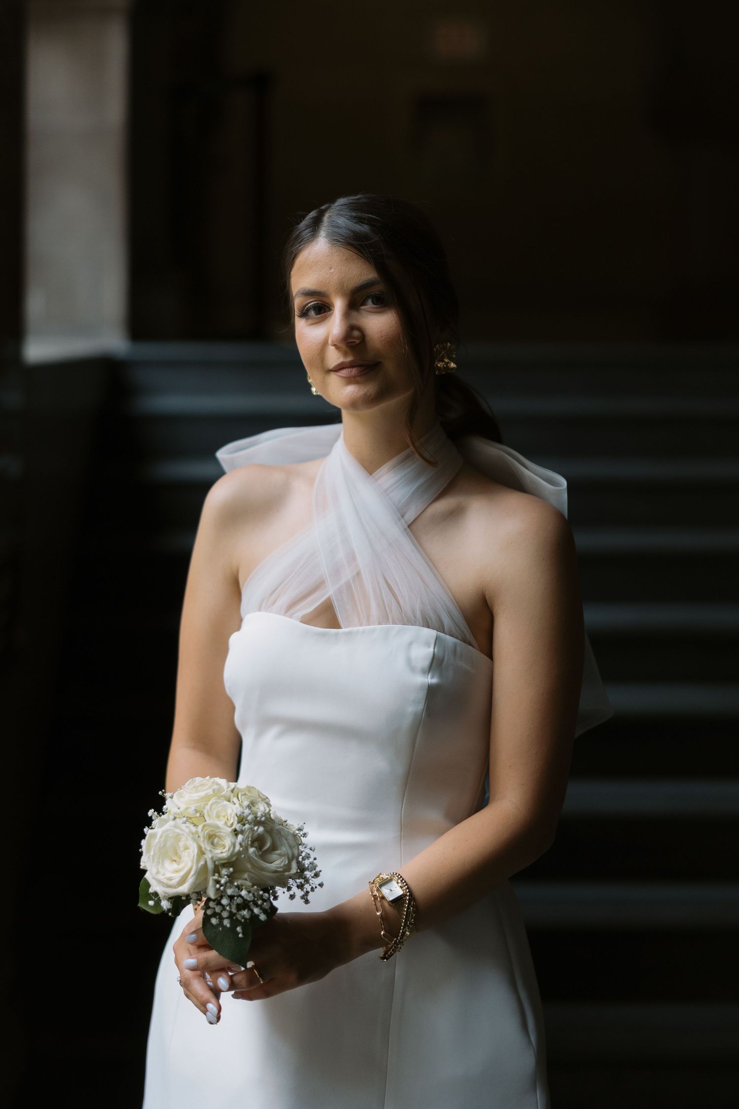 A bride in a white halter dress holds a bouquet of white roses while standing in a dimly lit hall.