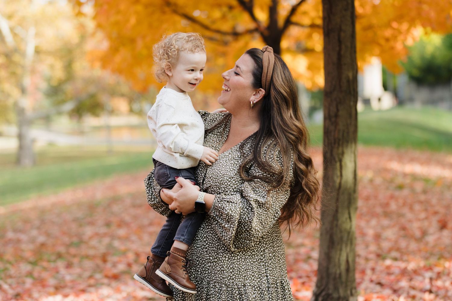 A joyful parent holds and laughs with a small child outdoors among vibrant autumn leaves and trees.