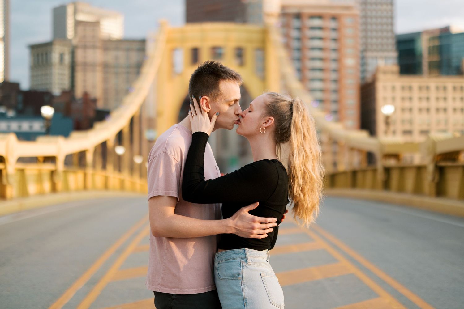 A romantic embrace on a golden bridge during sunset with city skyline in background.