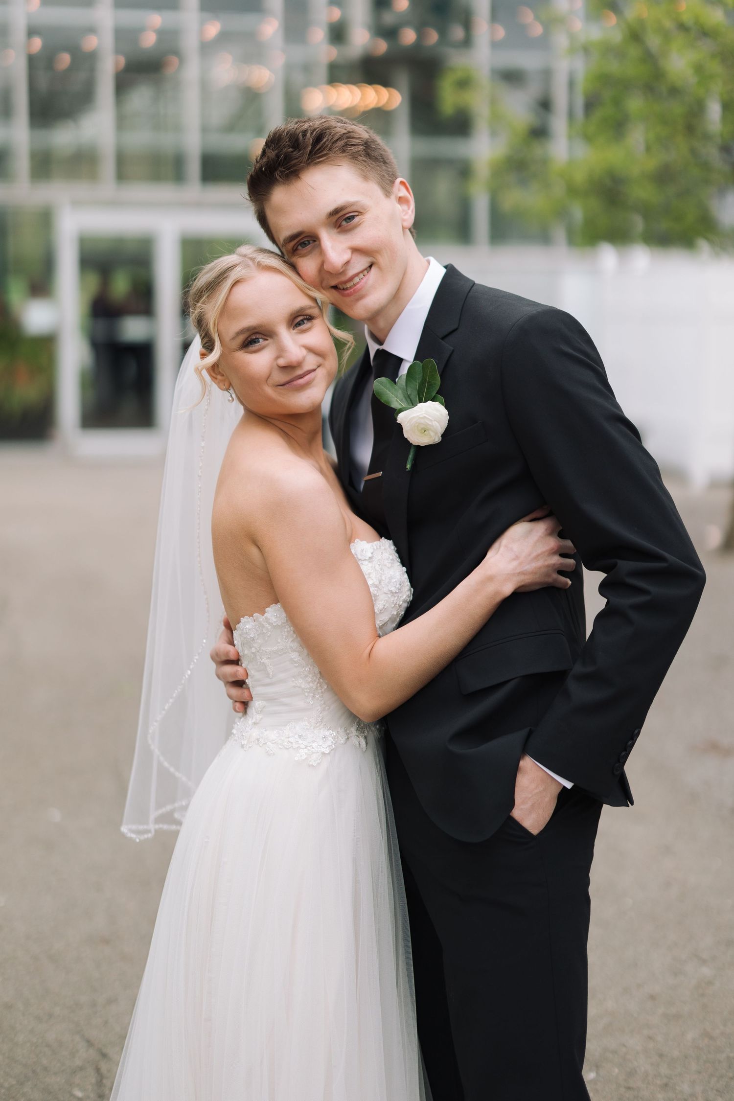 Smiling bride in white gown and veil embraces groom in black suit with white boutonniere outdoors.