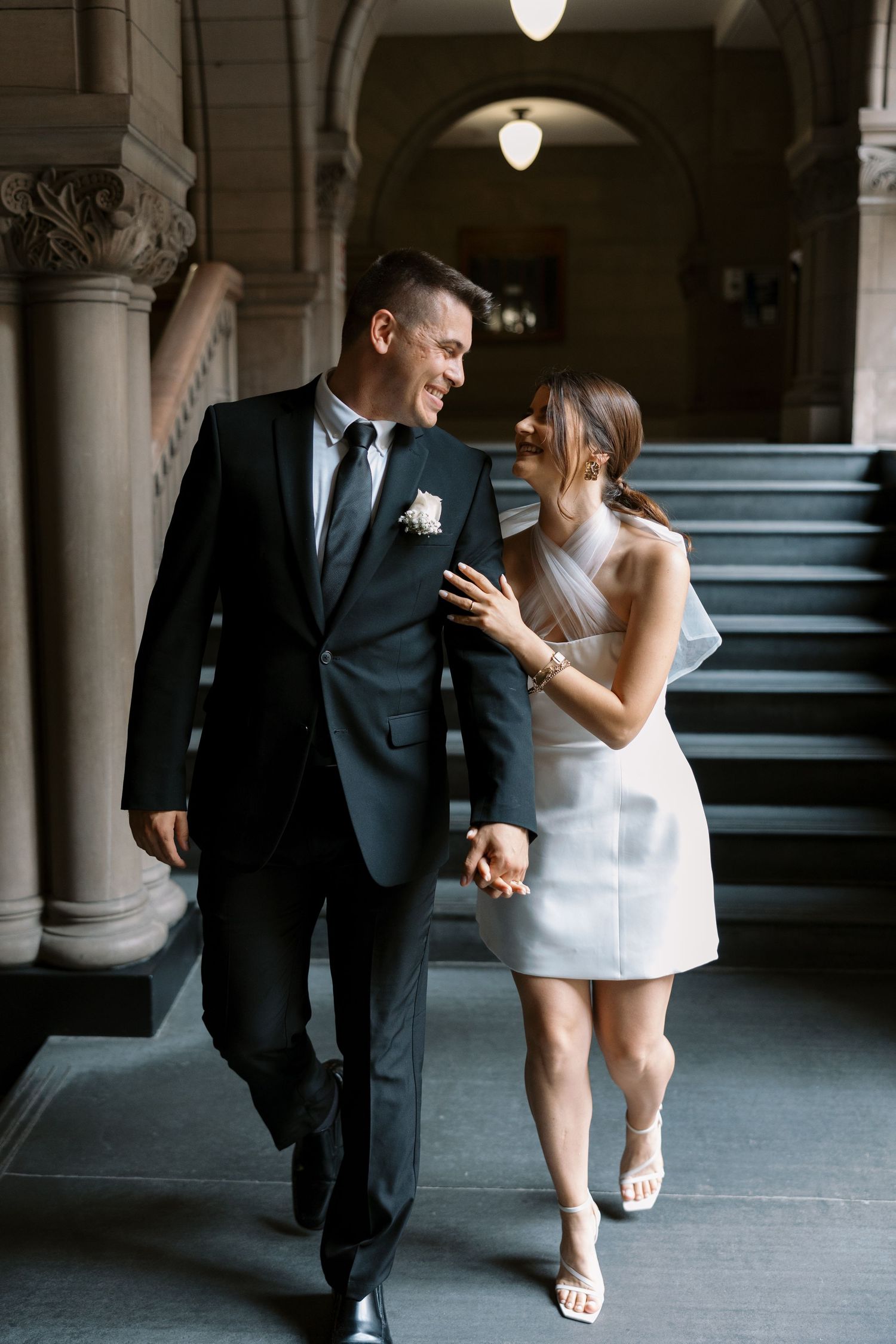 A couple walks together through an elegant hallway with stone columns and arched lighting.