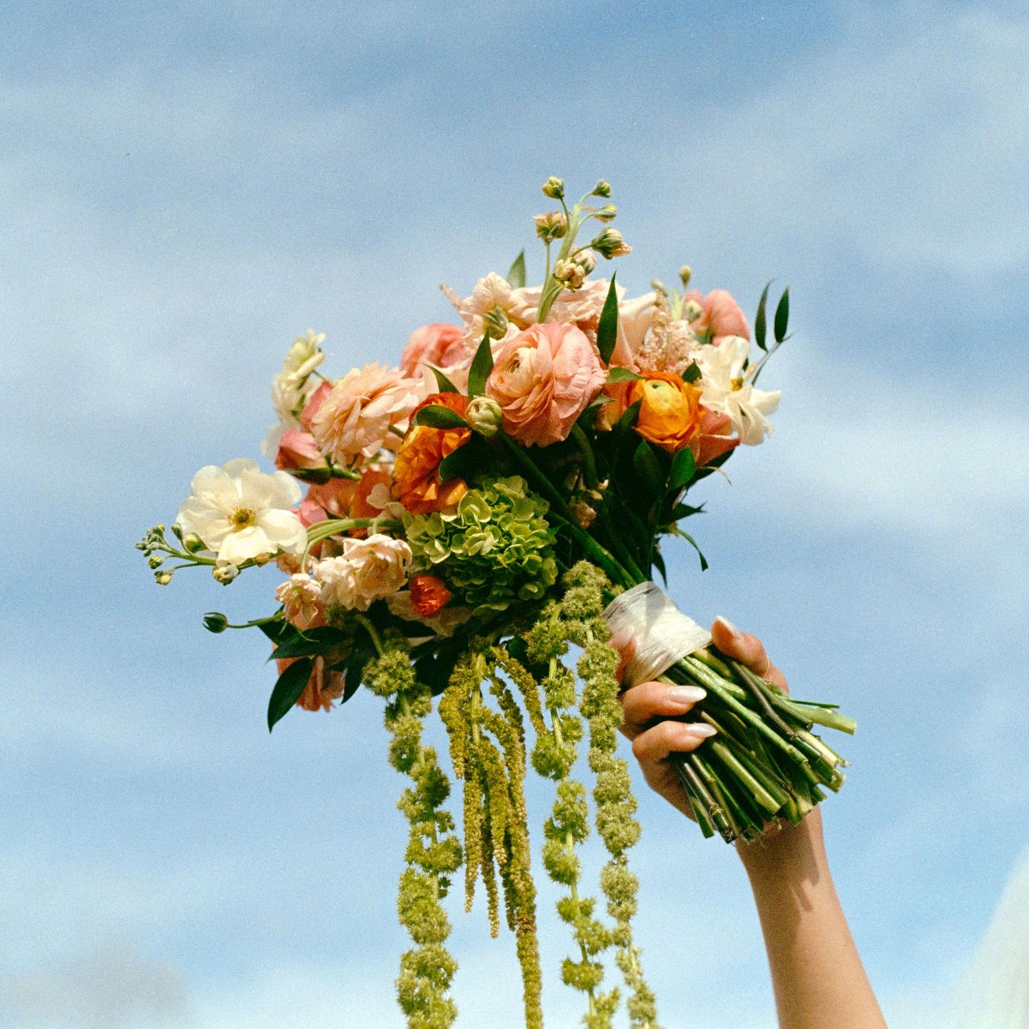 A hand holds a vibrant bridal bouquet of pink, orange, and white flowers against a blue sky.
