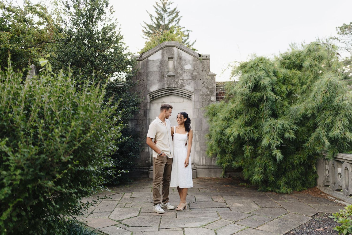 A couple walks along a stone path surrounded by lush greenery and gardens in a romantic outdoor setting.
