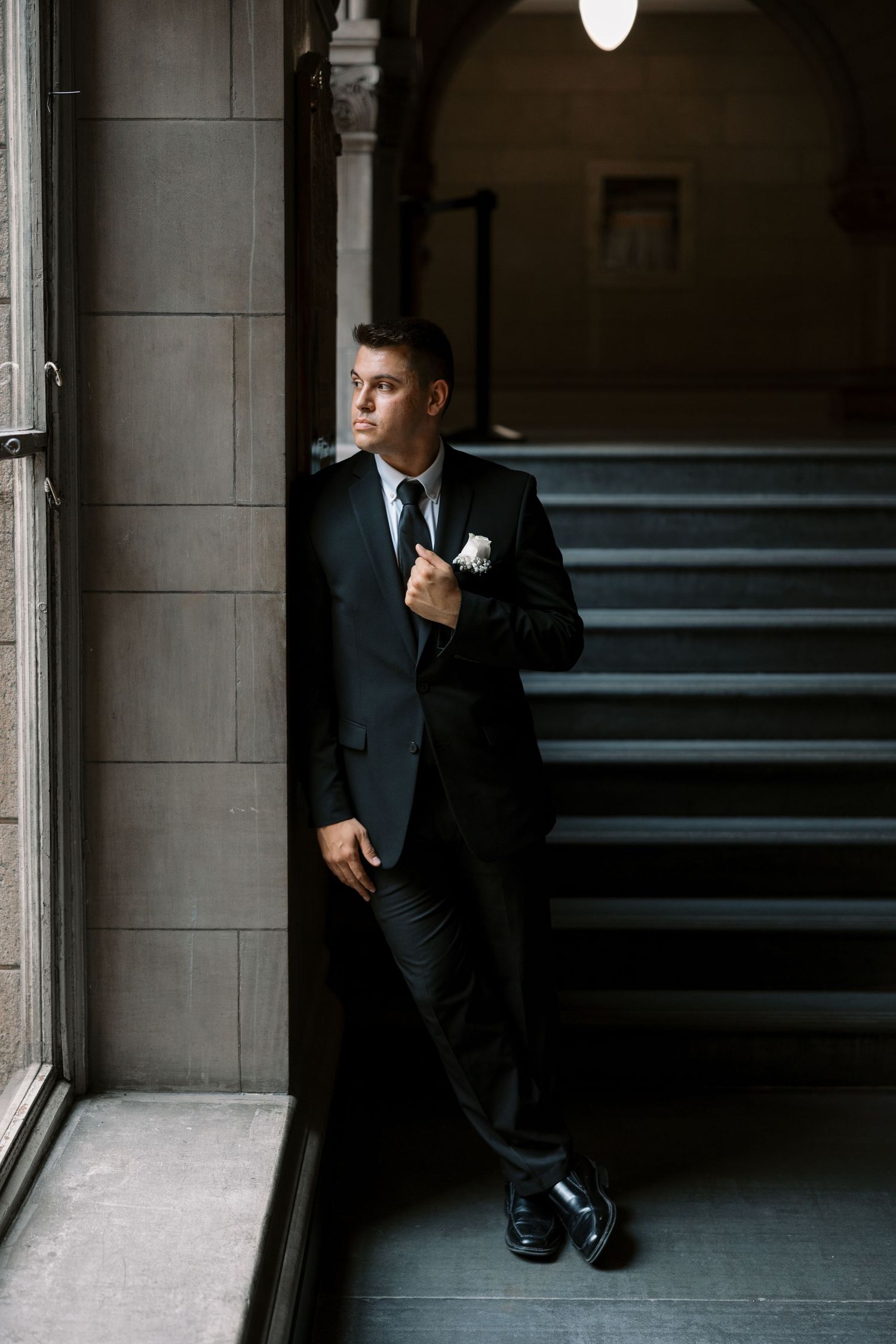 A groom in a classic black suit poses against a window in a historic building with stone architecture.