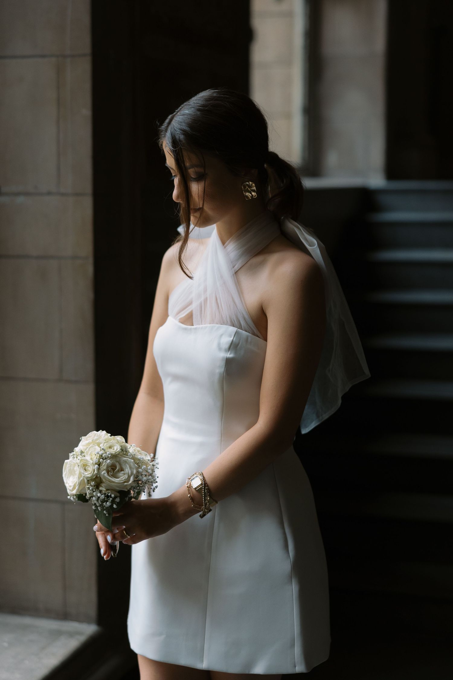 A bride in a short white dress holds a bouquet while standing in a vintage building.