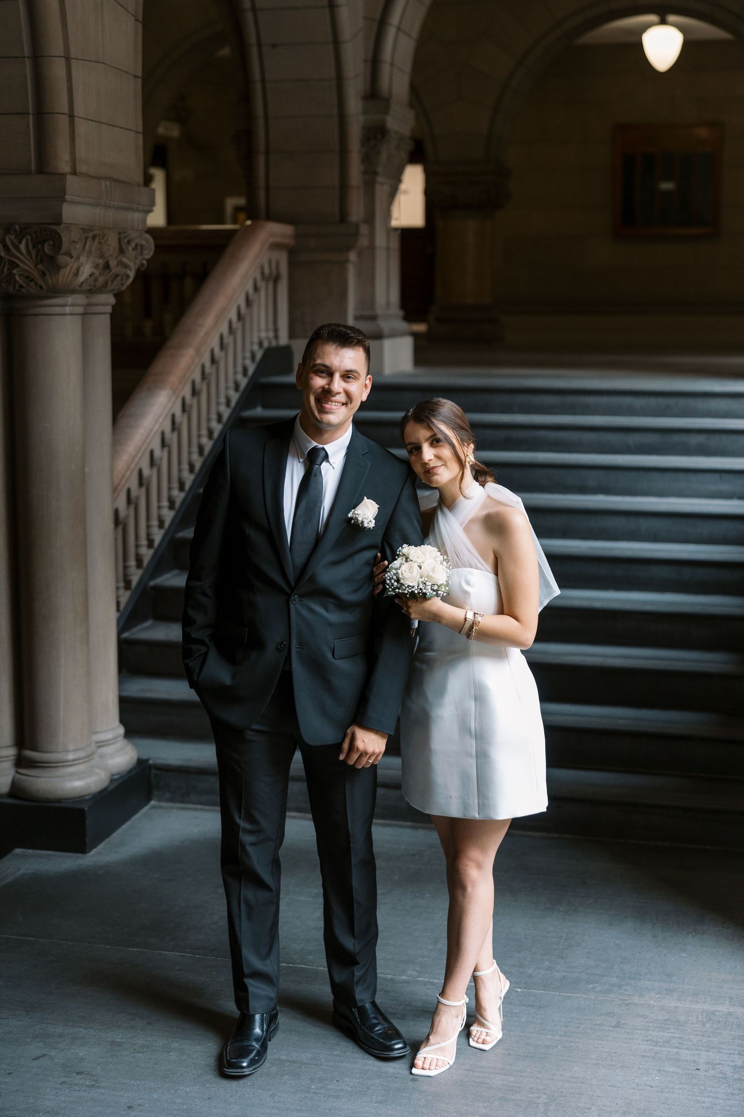 Elegant couple poses on historic stone steps in formal wedding attire with bouquet.