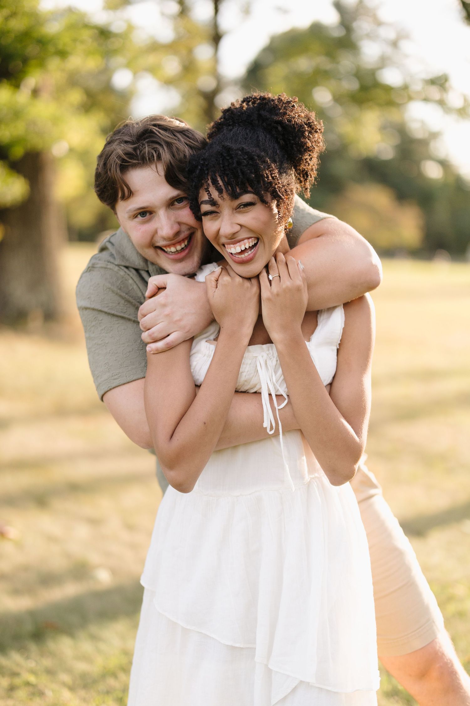 A couple shares a playful candid moment embracing and laughing together in a golden sunlit outdoor setting.