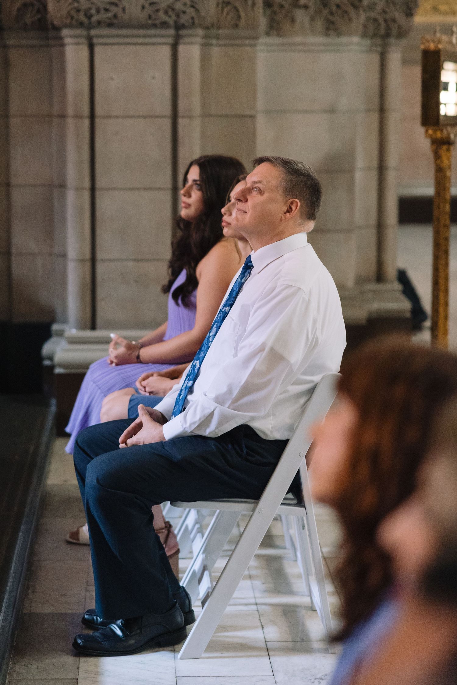Two guests sit on white chairs in an elegant hallway with stone arches and columns.