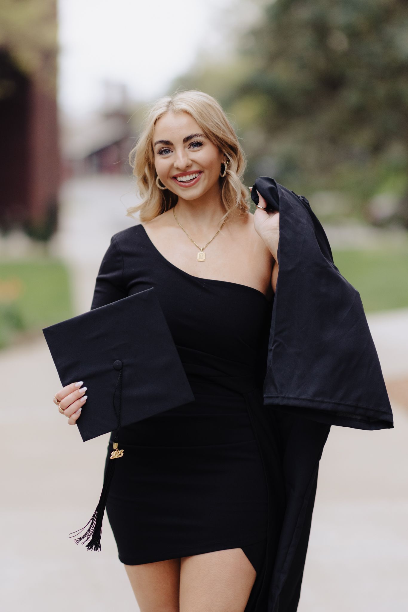 Graduate in black dress holds cap and gown while posing on campus pathway.
