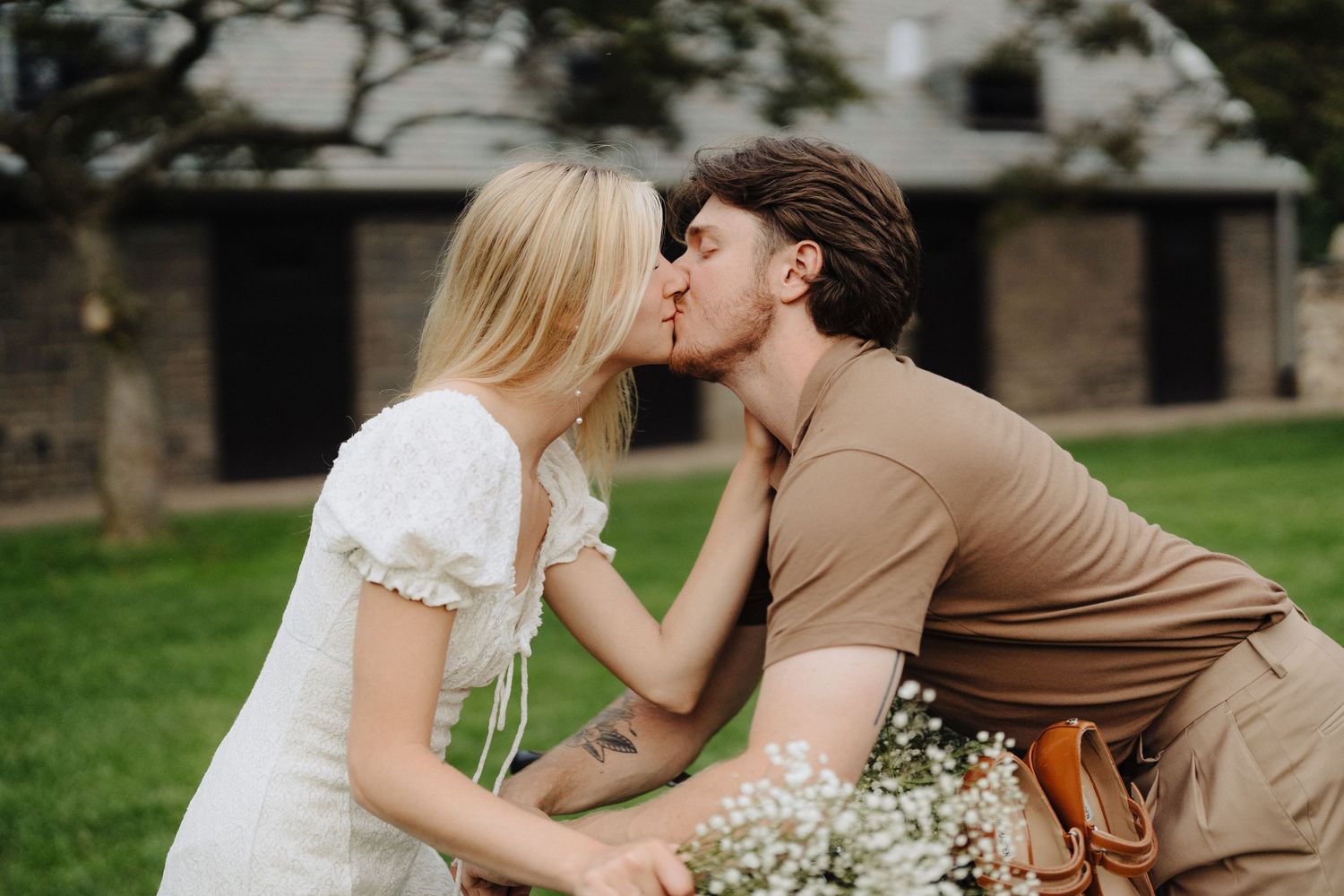 A tender moment shared on a bicycle against a pastoral backdrop with brick buildings.