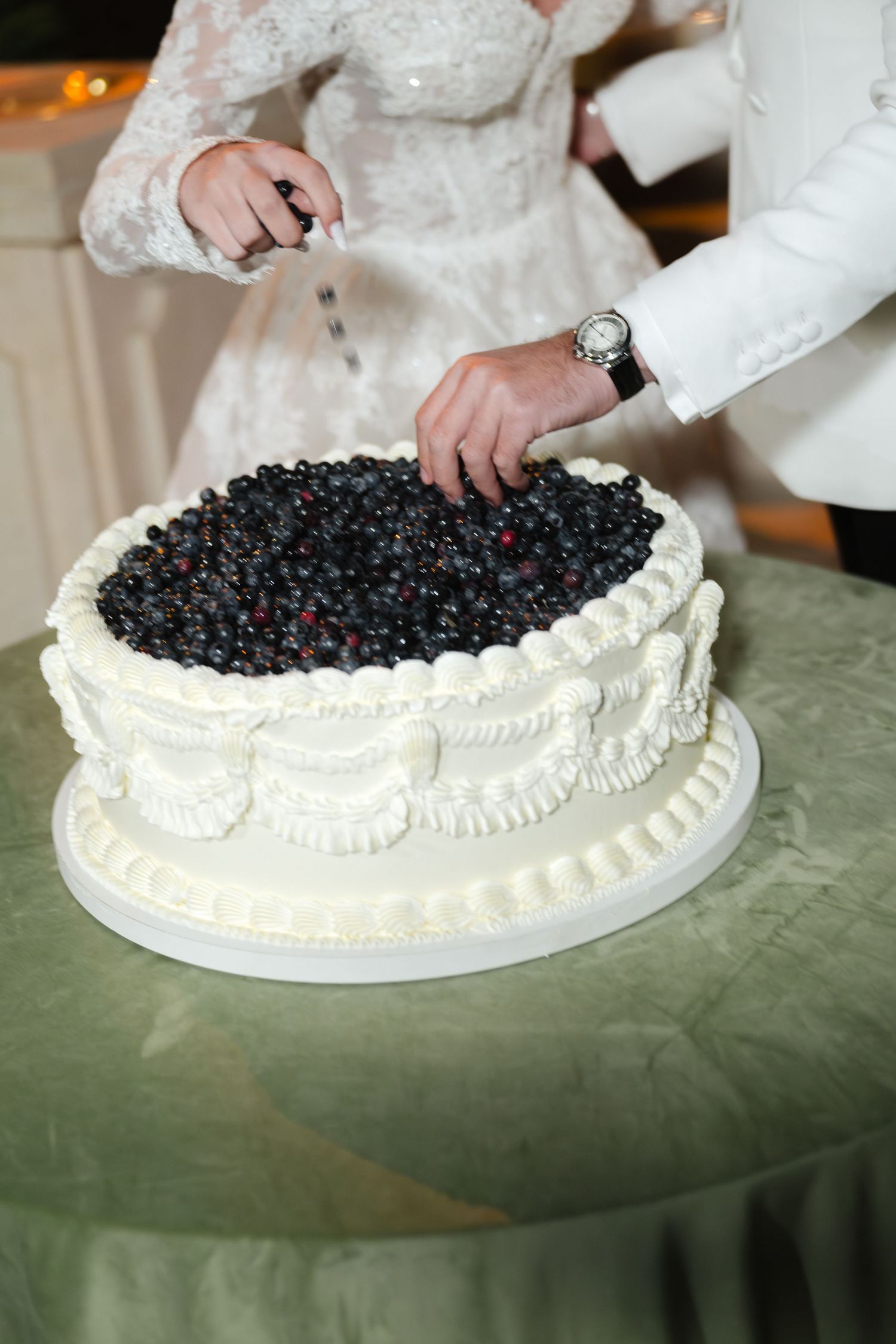 Bride and groom cutting a white wedding cake topped with fresh blueberries and elegant cream frosting details.