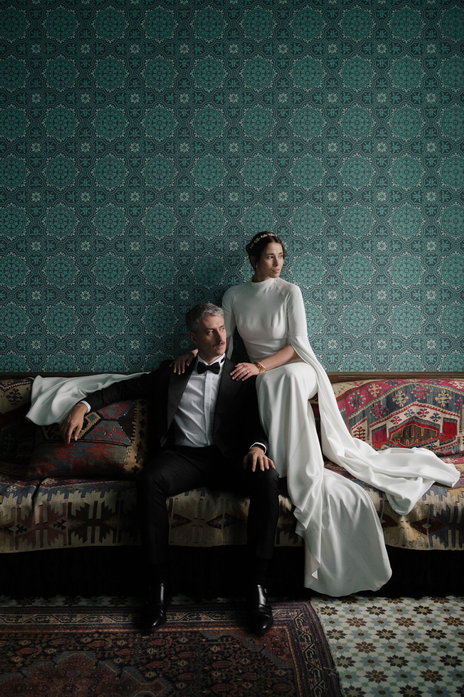Elegant bride in white gown and groom in tuxedo posing on vintage sofa against teal patterned wallpaper.
