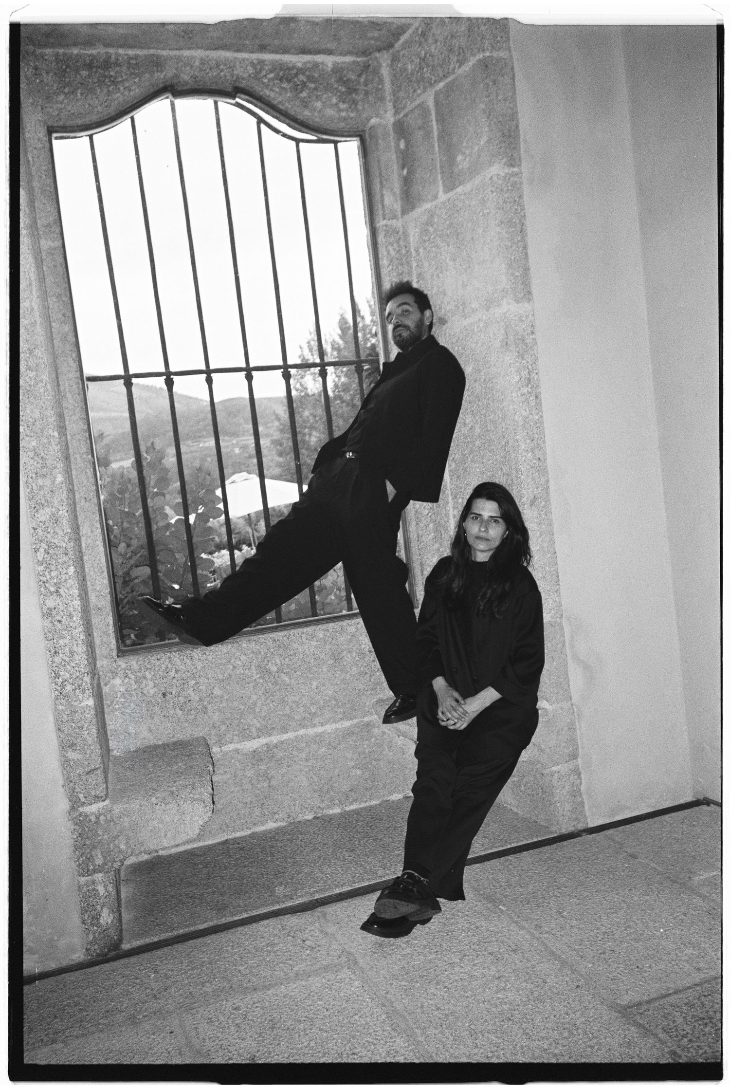 Black and white photo of two people dressed in black posing by a stone archway with iron bars.