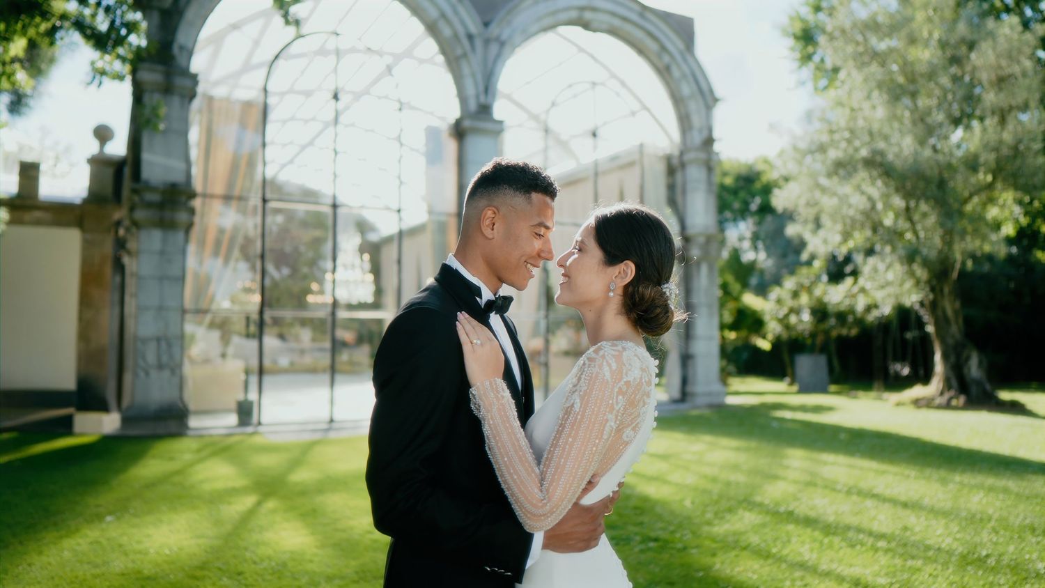 A couple shares romantic moments in a greenhouse garden setting with natural sunlight streaming through glass panels.