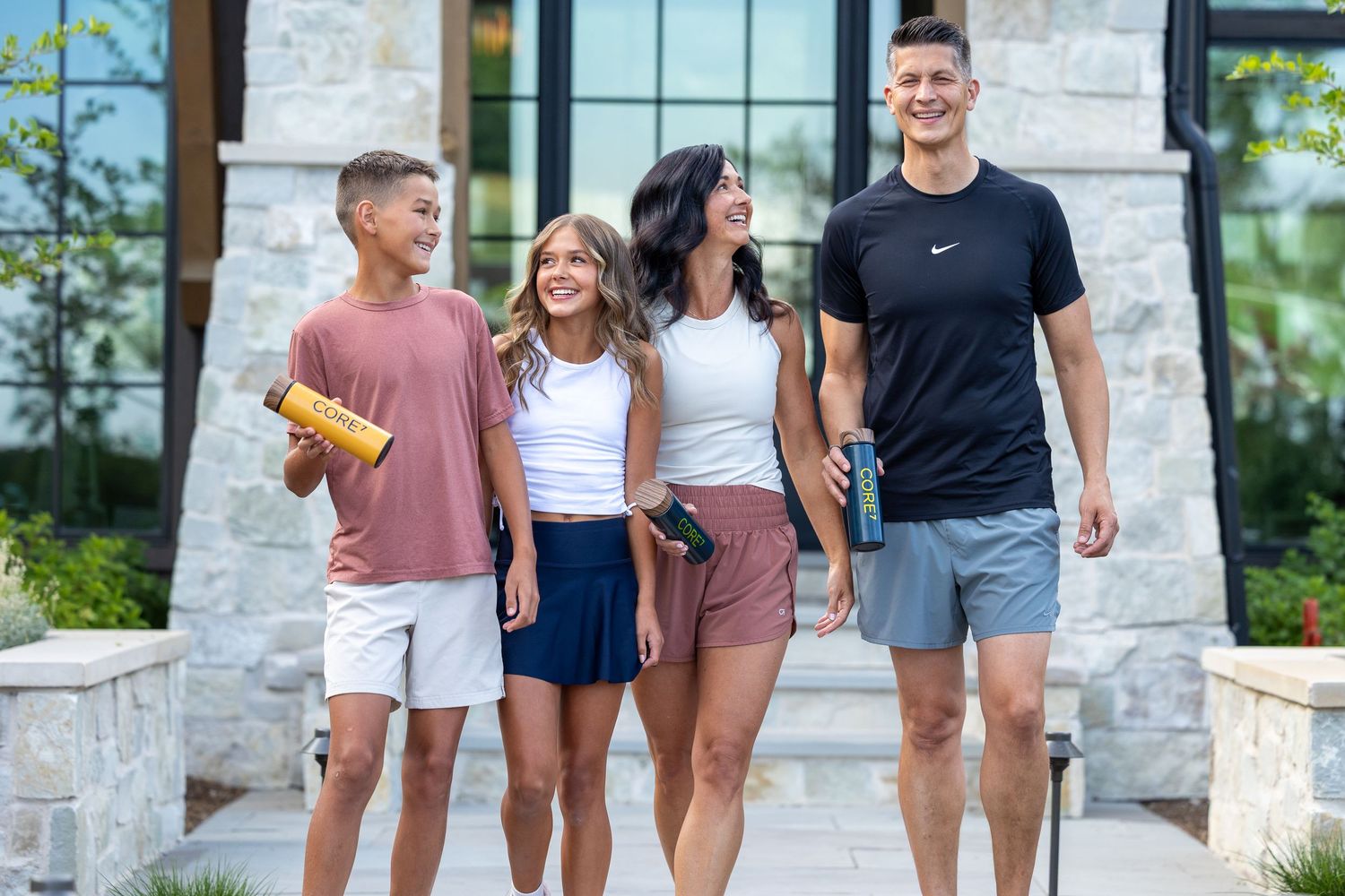 Happy family of four walking together outdoors holding water bottles on a sunny day.