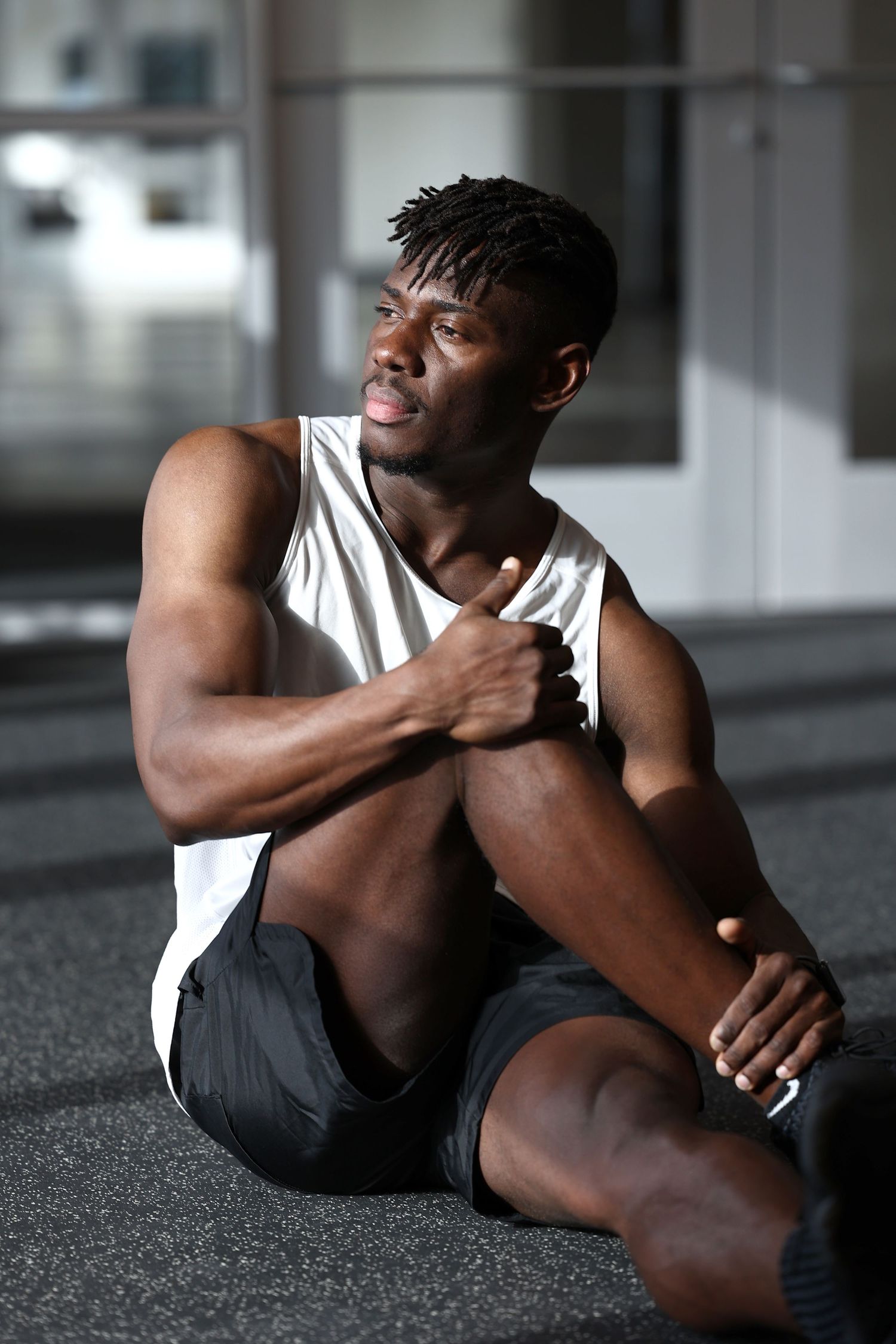 Muscular Black man in white tank top resting on gym floor after workout.