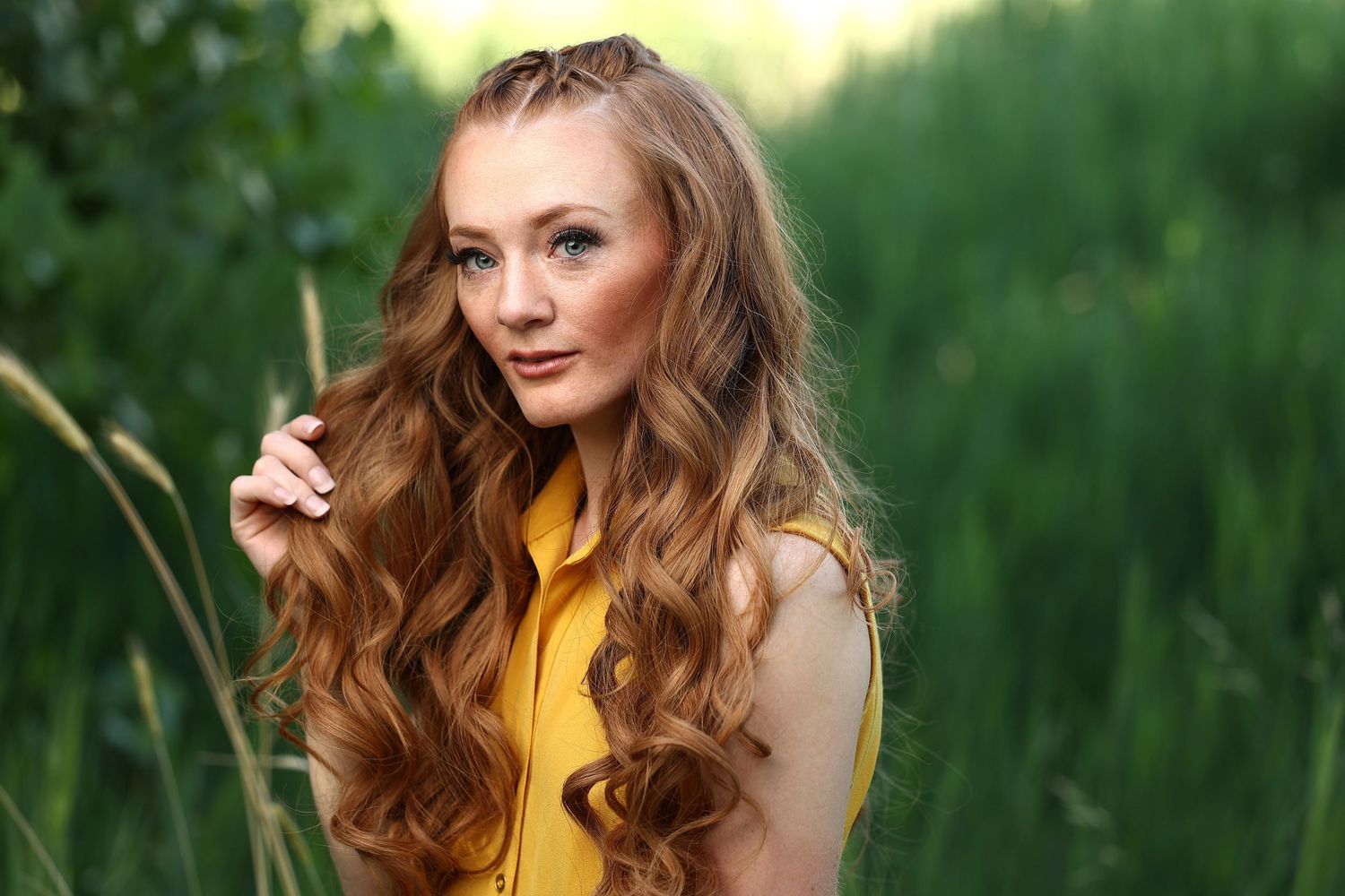 Young woman with long curly auburn hair posing outdoors in a yellow dress among green foliage.