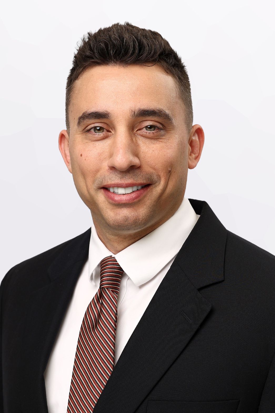 Professional headshot of a confident man in a black blazer and striped tie with a light background.