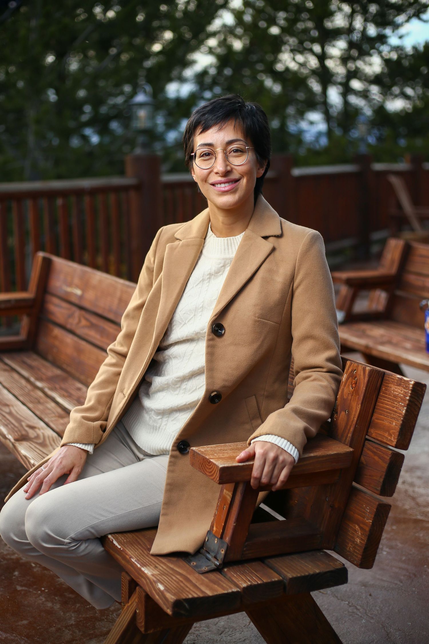 Woman in beige blazer sitting on wooden bench outdoors smiling wearing glasses.