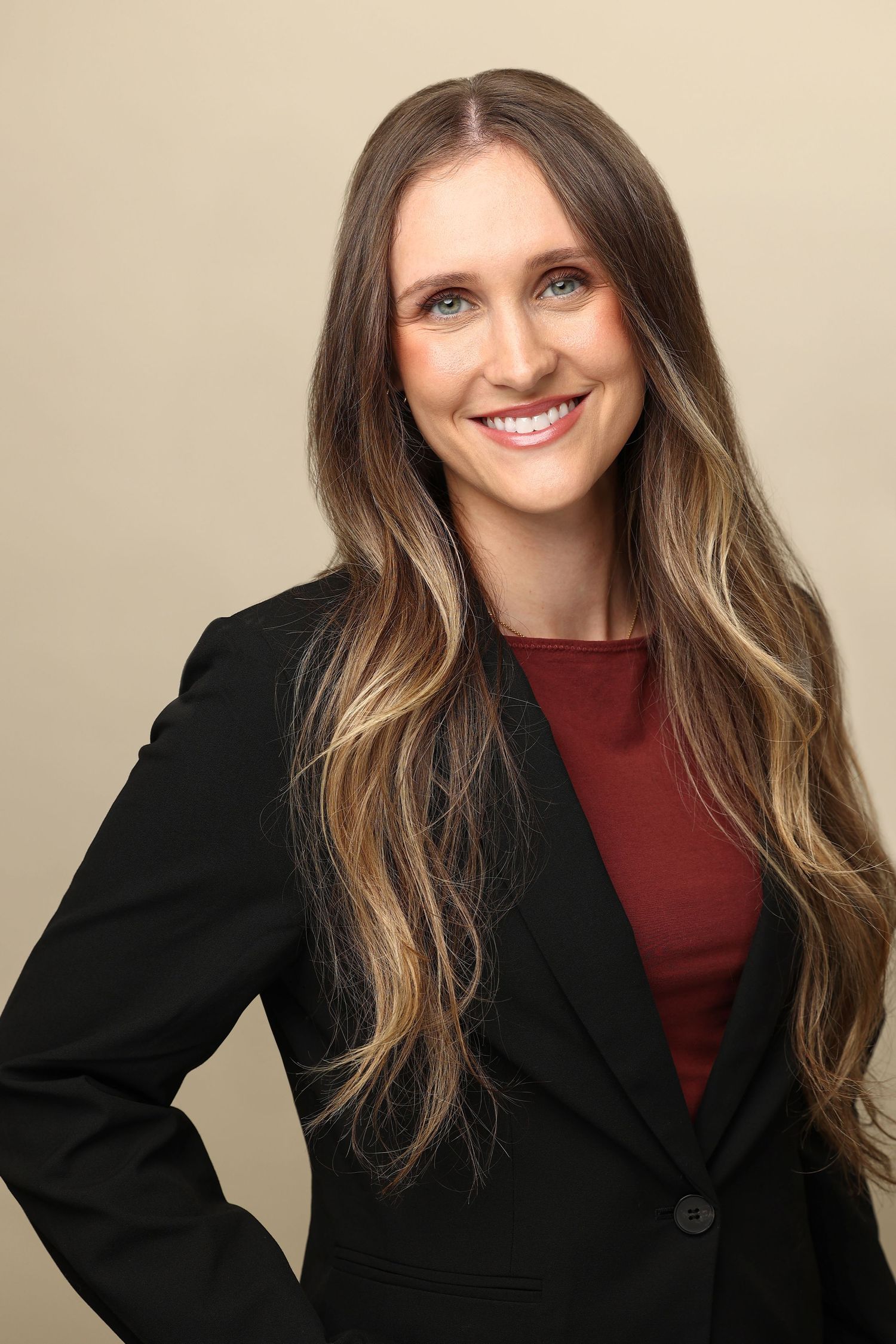 Professional headshot of a smiling woman with long highlighted hair wearing a black blazer and red top.