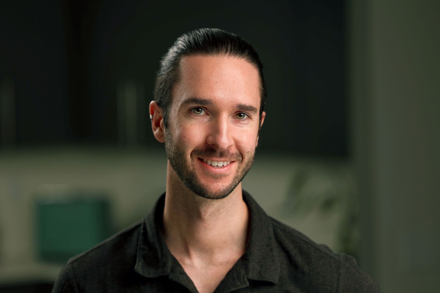 Smiling young man with dark hair wearing a black shirt in a softly blurred indoor setting.